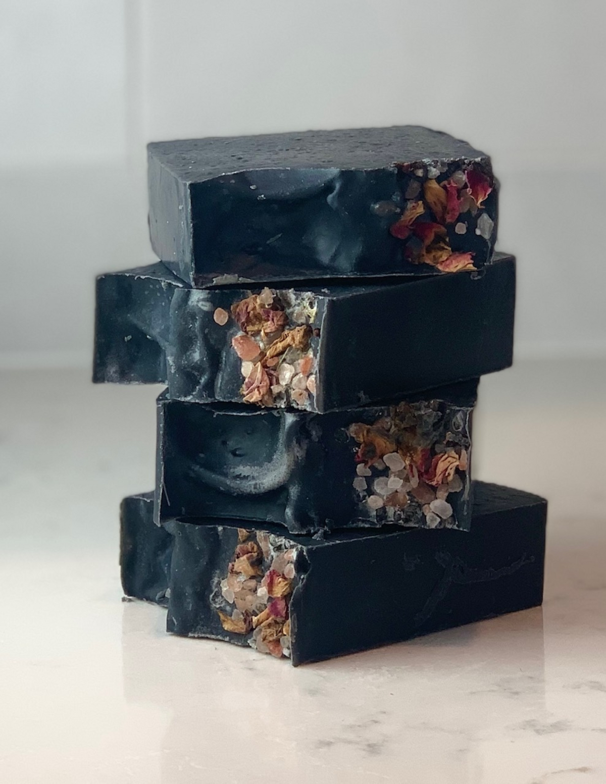 Stack of four black color charcoal and tea tree essential oil soap on a white marble table. Adorned with pink sea salt on top for decoration. 
