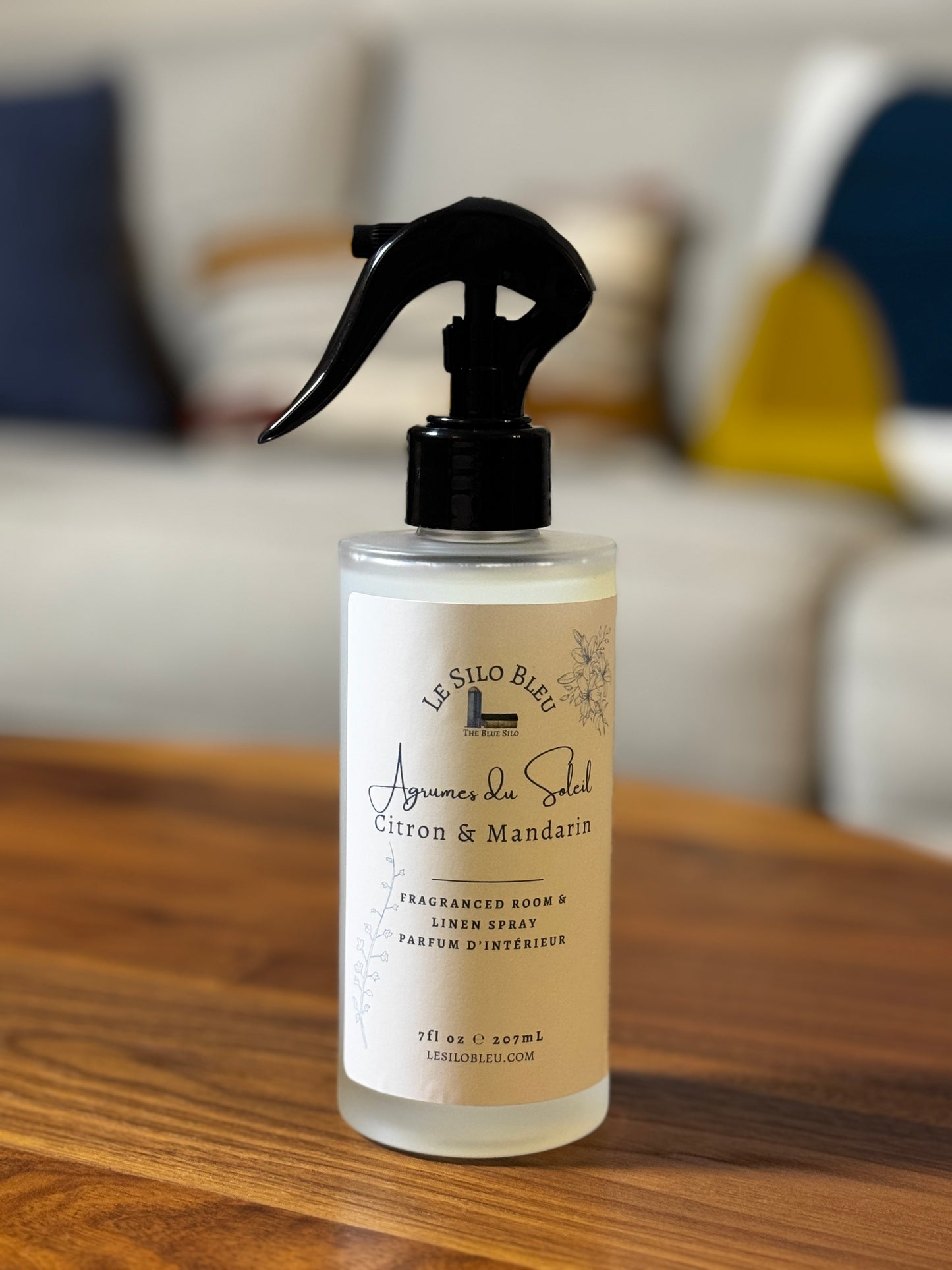 A bottle of La Silo Bleu Citron & Mandarin room spray, featuring a frosted bottle with a black trigger sprayer and a cream-colored label, set on a wooden table with a blurred living room background.