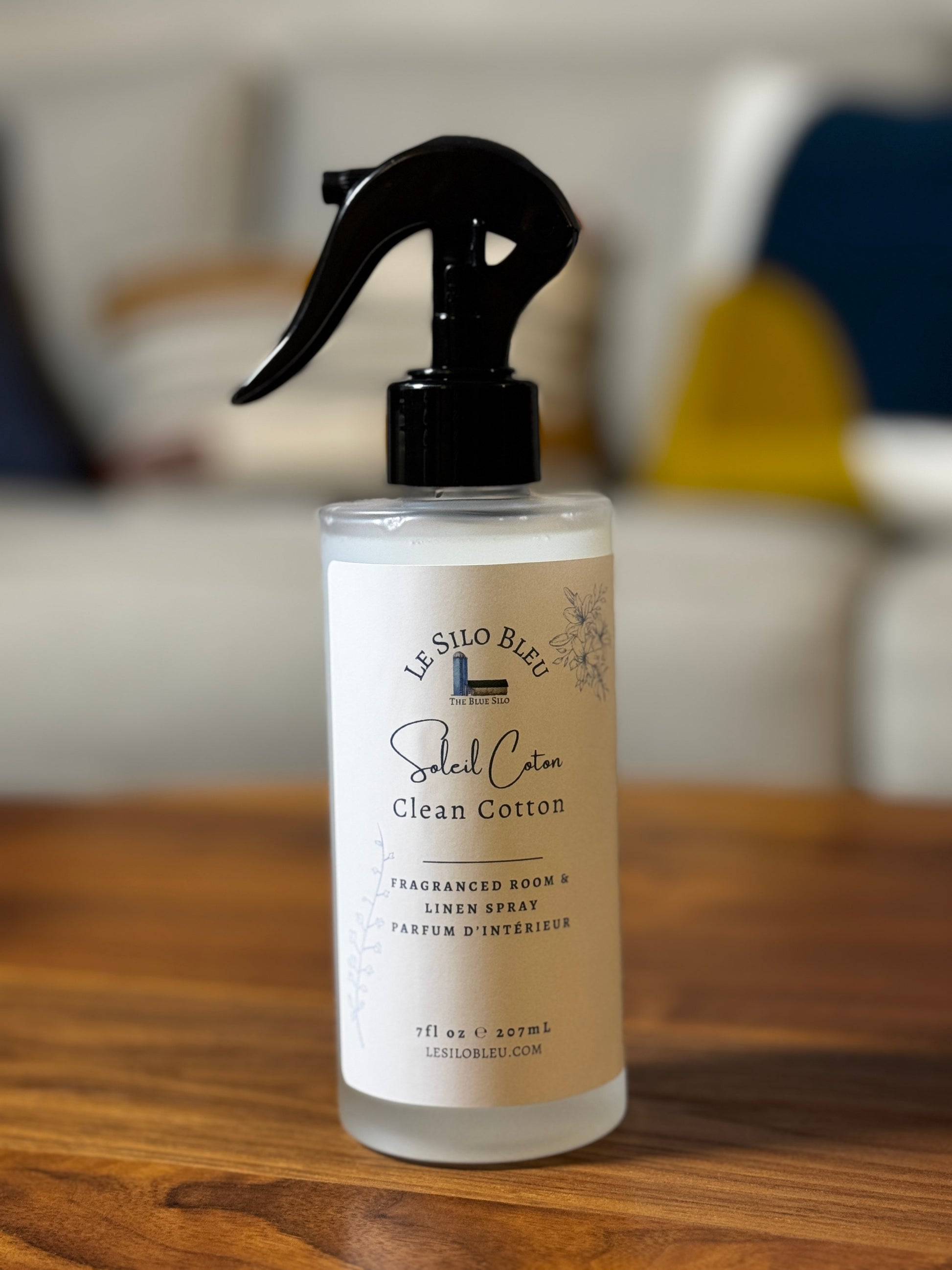 A bottle of Le Silo Bleu Clean Cotton Room & Linen Spray, featuring a black trigger spray top and a cream-colored label, set on a wooden table with a blurred home background.