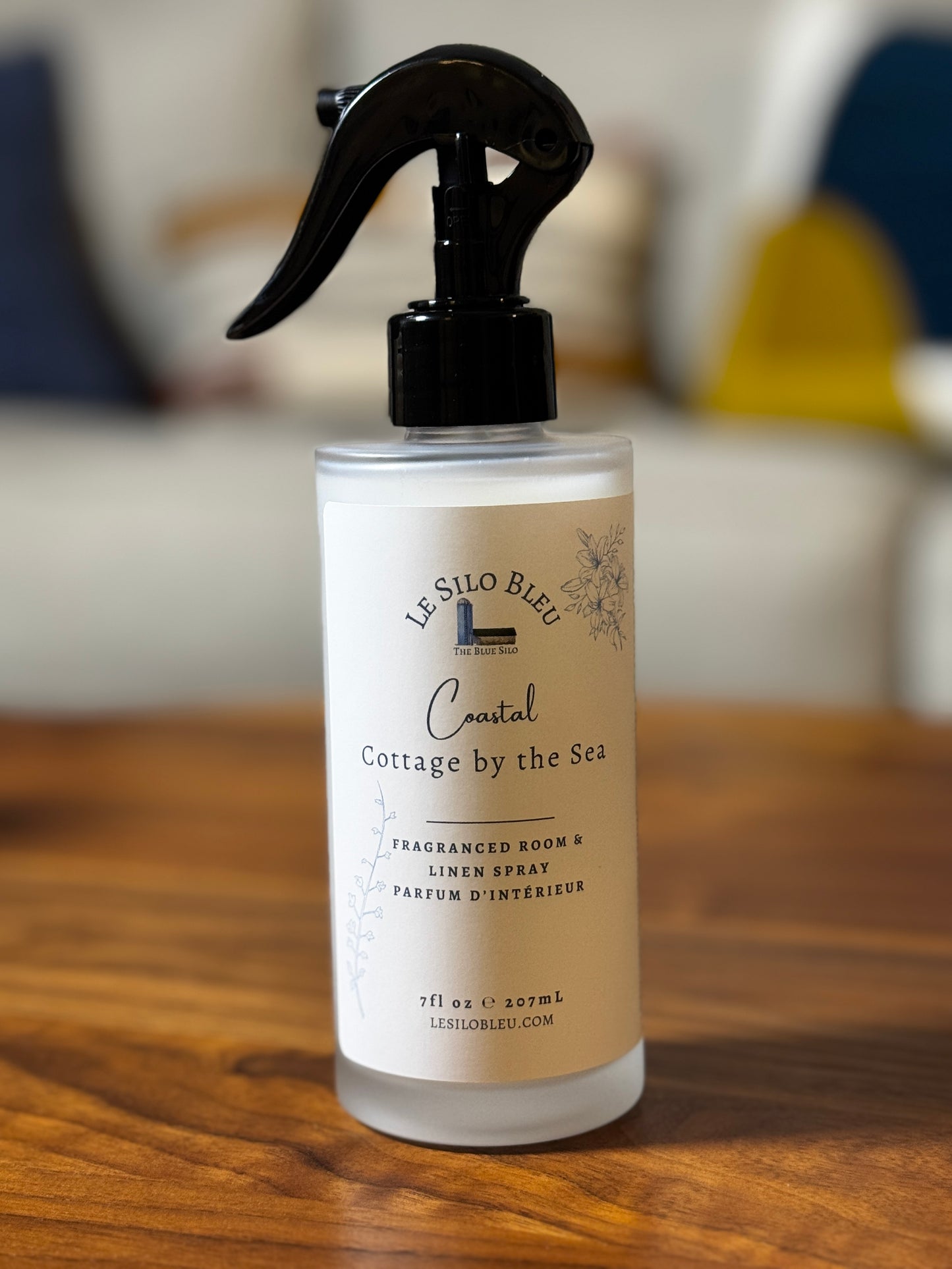 A frosted glass spray bottle of Le Silo Bleu Coastal Cottage by the Sea Fraganced Room & Linen Spray, featuring a black trigger sprayer and a cream-colored label, set on a wooden table with a blurred background.