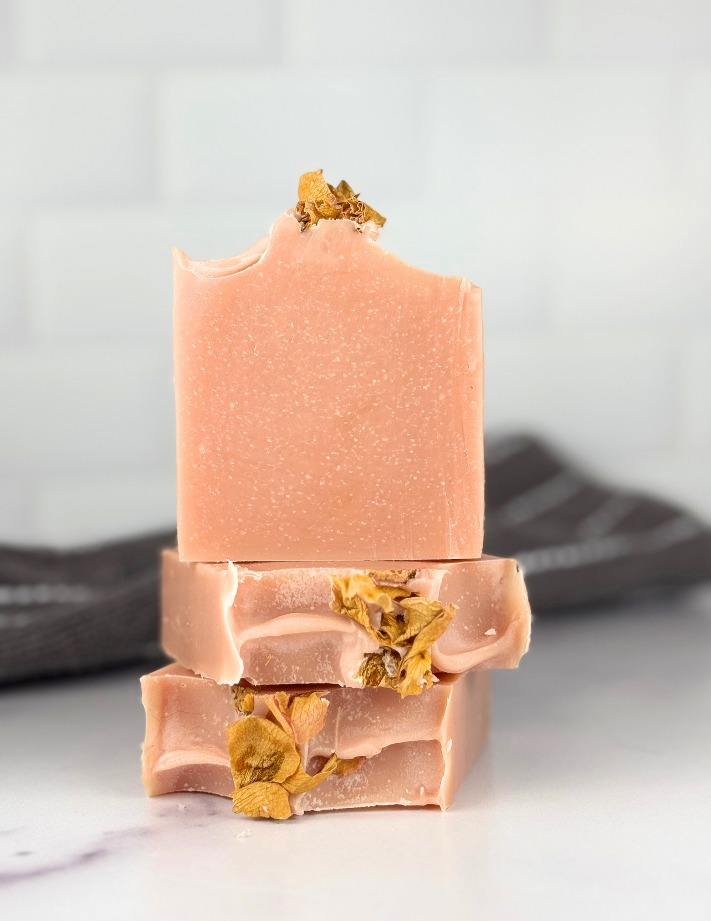 Three bars of pink soap with rose petals on top sitting on a white marble table with a grey and white stripped towel in the background. 