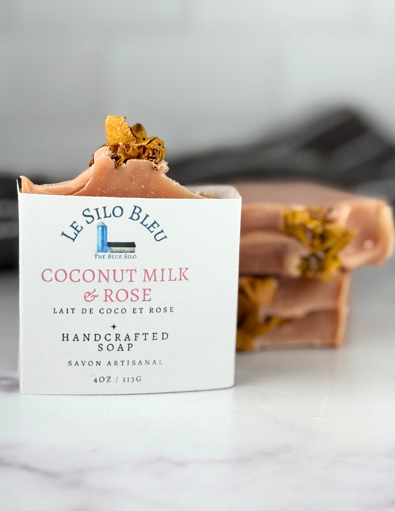 One labeled bar and two naked bars of pink soap with rose petals on top sitting on a white marble table with a grey and white stripped towel in the background. 