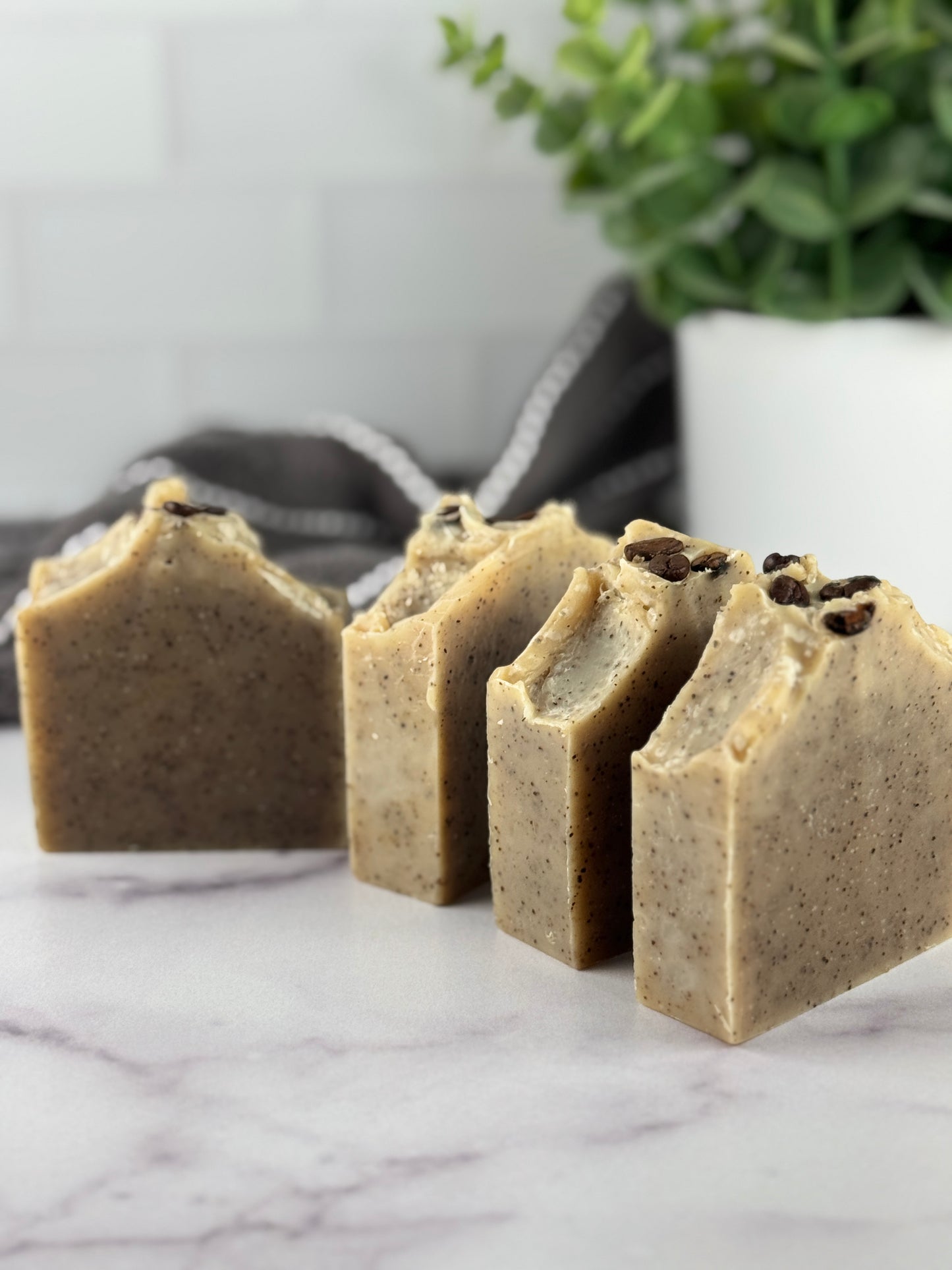 Four soap bars with visible coffee beans on a marble surface, with a plant in the background.