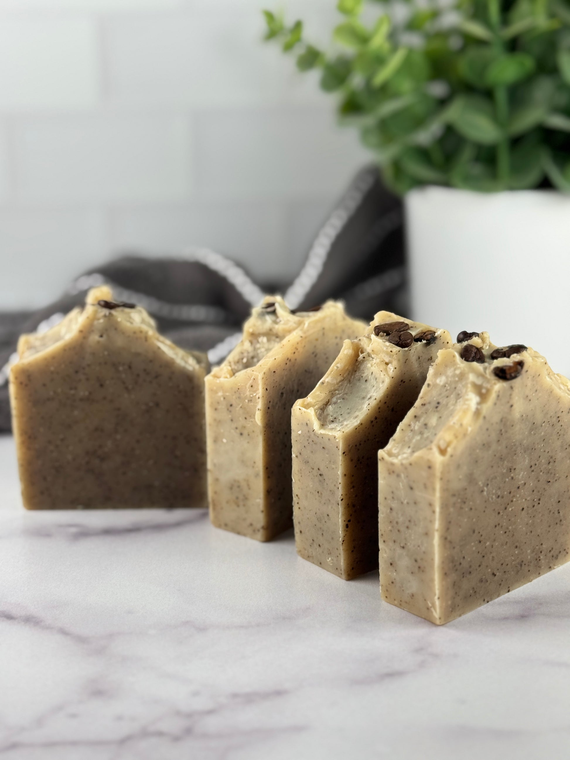 Four soap bars with visible coffee beans on a marble surface, with a plant in the background.