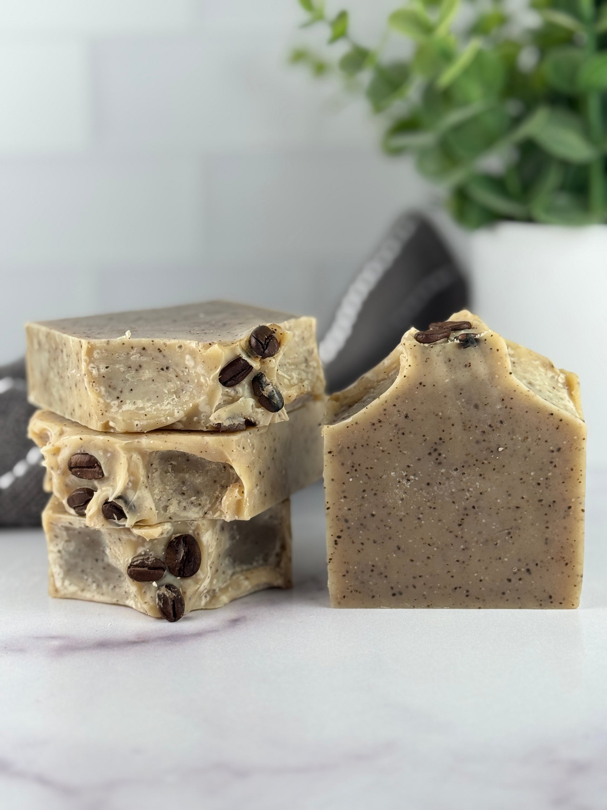 Three bars of soap with coffee beans on a marble surface, with a plant in the background.