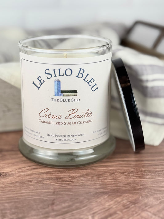 Large scented soy wax candle in jar with the lid to the side, with the label 'LE SILO BLEU' and 'CÉme Brülée Caramelized Sugar Custard'.  The jar is sitting on a wooden table with a basket with a dish cloth in the background.
