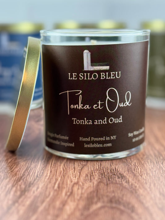 Tonka and Oud Scented Soy Wax Candle with 'Le Silo Bleu' label on a wooden surface with a gold lid leaning against the jar.