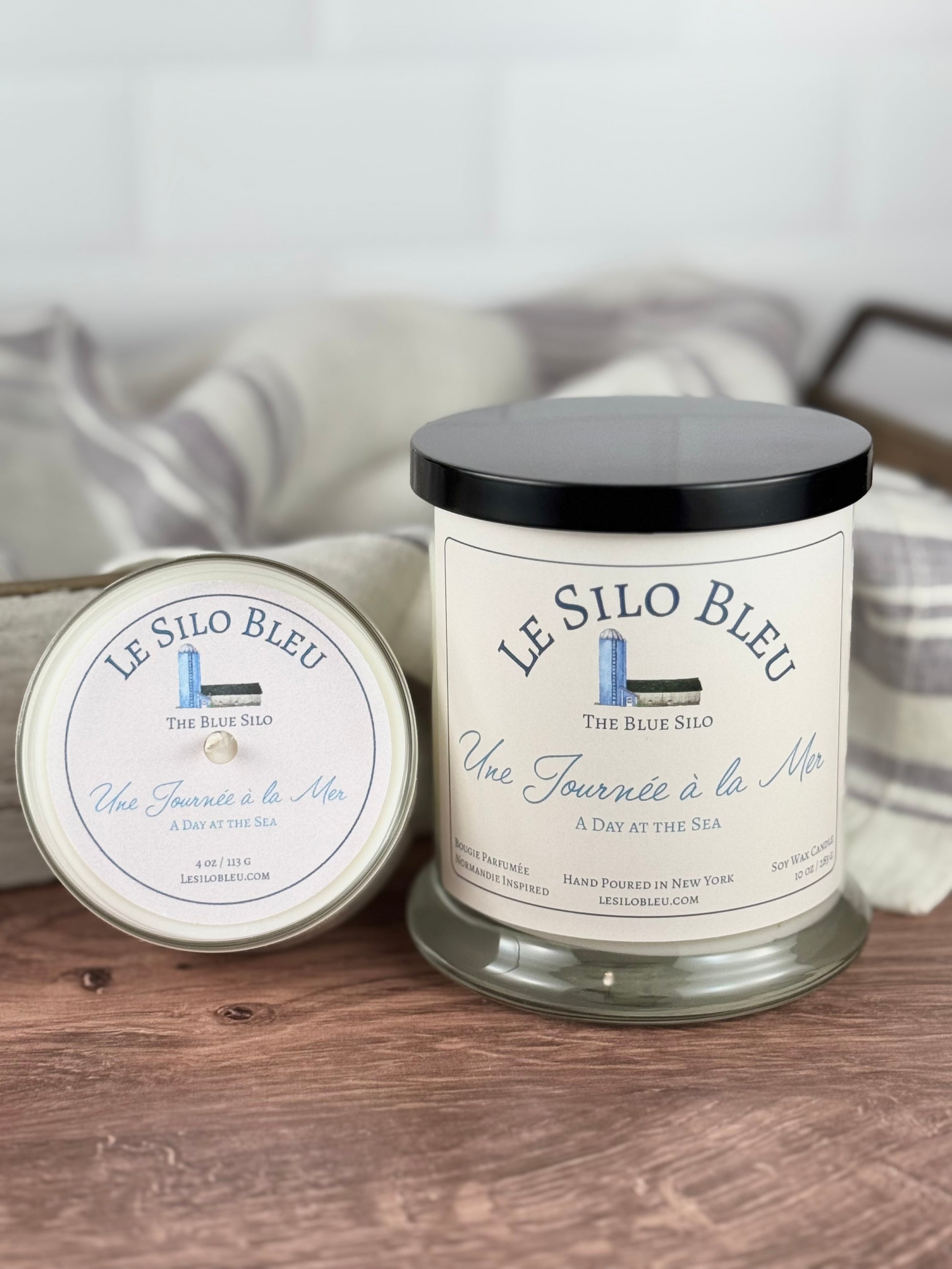 A large and small handpoured unlit soy wax candle in glass jars labeled "Une Journee a la Mer- Day at the Sea" sitting on a wooden table top with a black lid on the jar. In the background there is a white linen towel with lavender stripes in an antique box with a metal handle. 