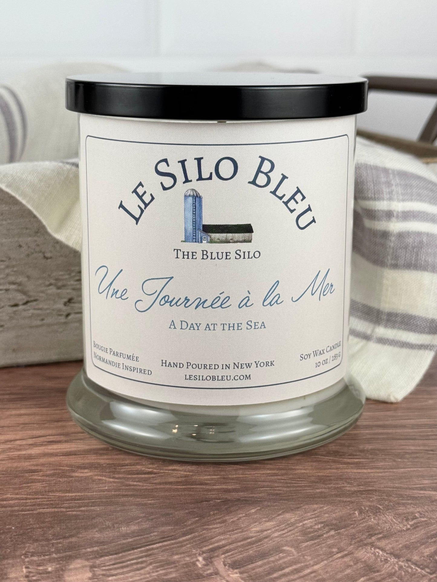 A large handpoured unlit soy wax candle in a glass jar labeled "Une Journee a la Mer- Day at the Sea" sitting on a wooden table top with a black lid on the jar. In the background there is a white linen towel with lavender stripes in an antique box with a metal handle. 