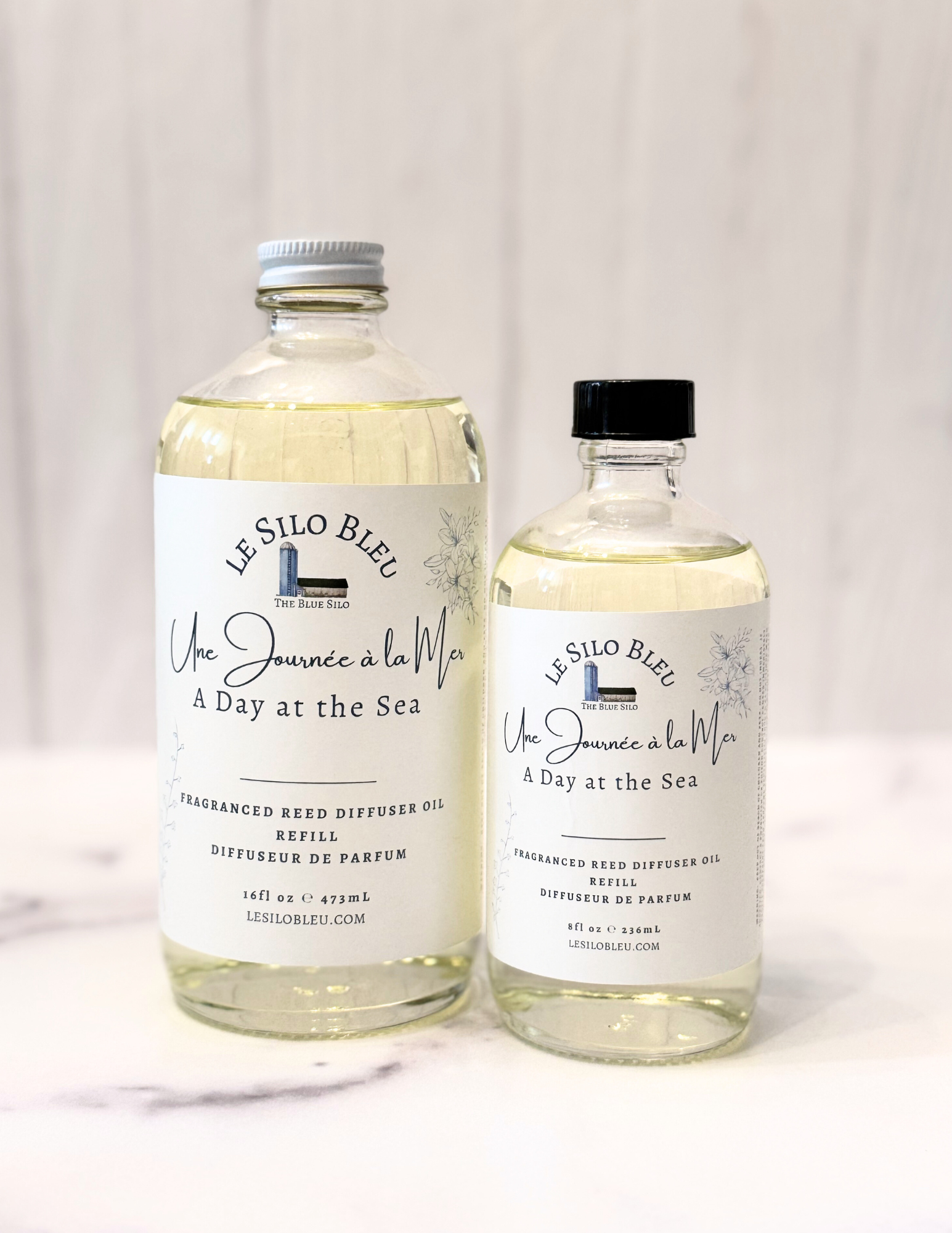 Day at the Sea Reed Diffuser Refill bottles in 8oz and 16oz glass bottles are shown sitting on a white marble table with a white wood background. The cap for the large bottle is white and the cap for the small bottle is black. 