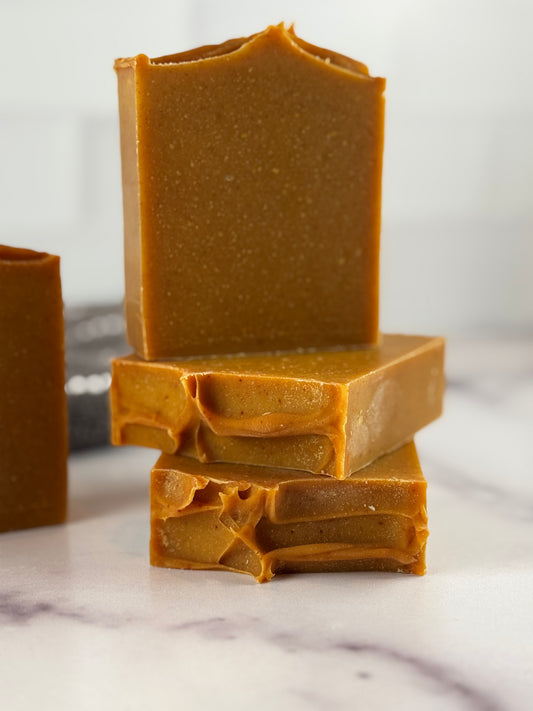 Three stacked bars of handmade soap in a deep turmeric color, photographed on a white marble surface with a soft-focus background.