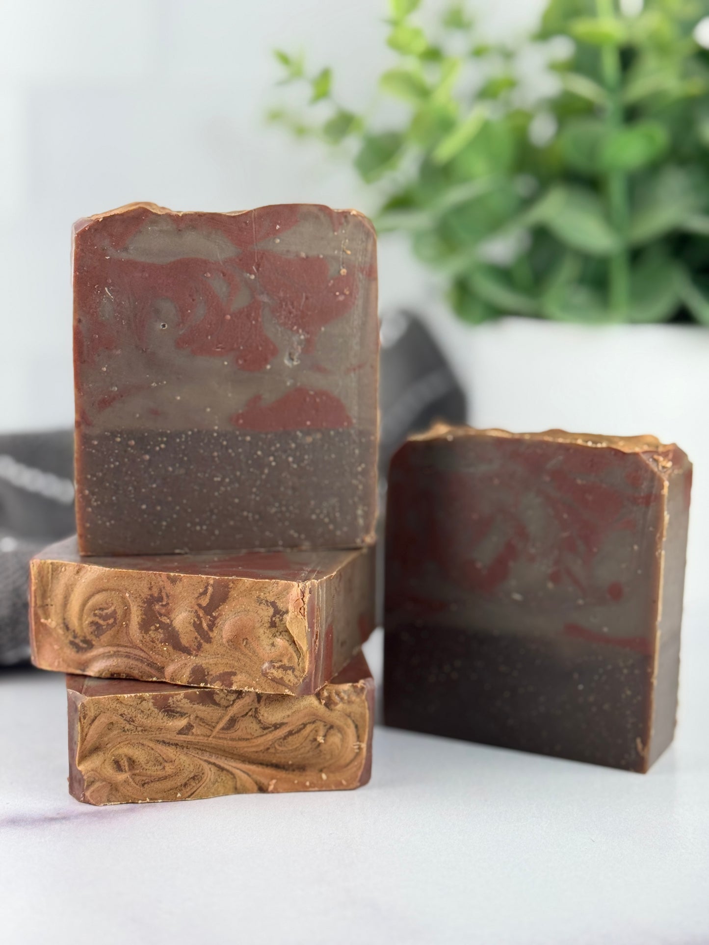 Three bars of brown soap with marbled patterns on a white surface with a blurred green plant in the background.