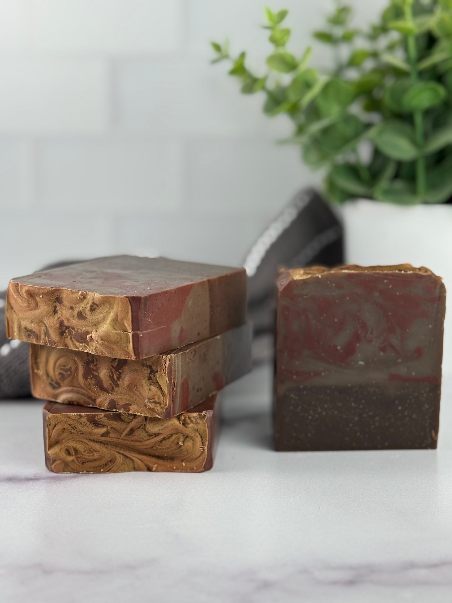 Three bars of soap with different textures and red brown and black colors on a marble surface with a plant in the background.