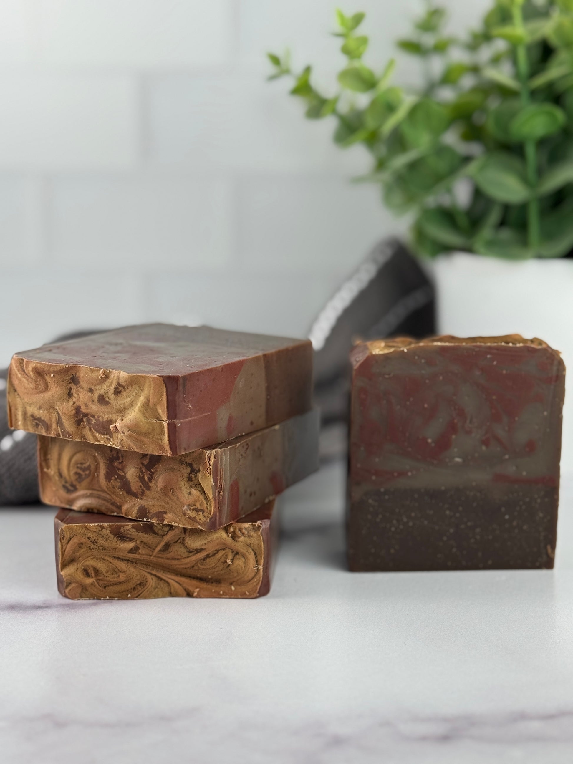 Three bars of soap with different textures and red brown and black colors on a marble surface with a plant in the background.