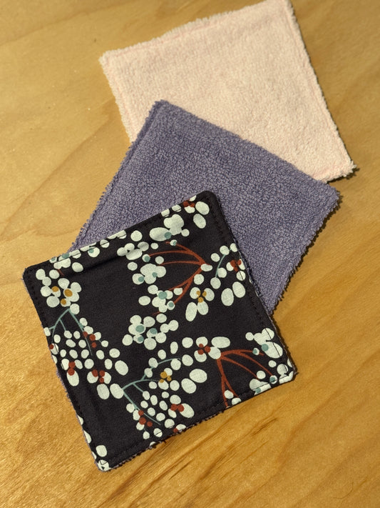 Three fabric face wash squares on a wooden surface:  dark purple with floral pattern, light pink and purple.