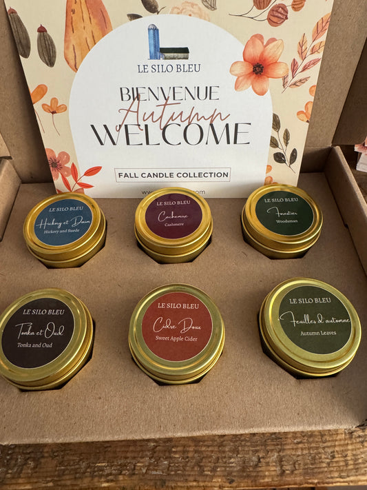 Set of six small candles in gold tins with visible labels on a cardboard box, featuring a 'Welcome' card.
