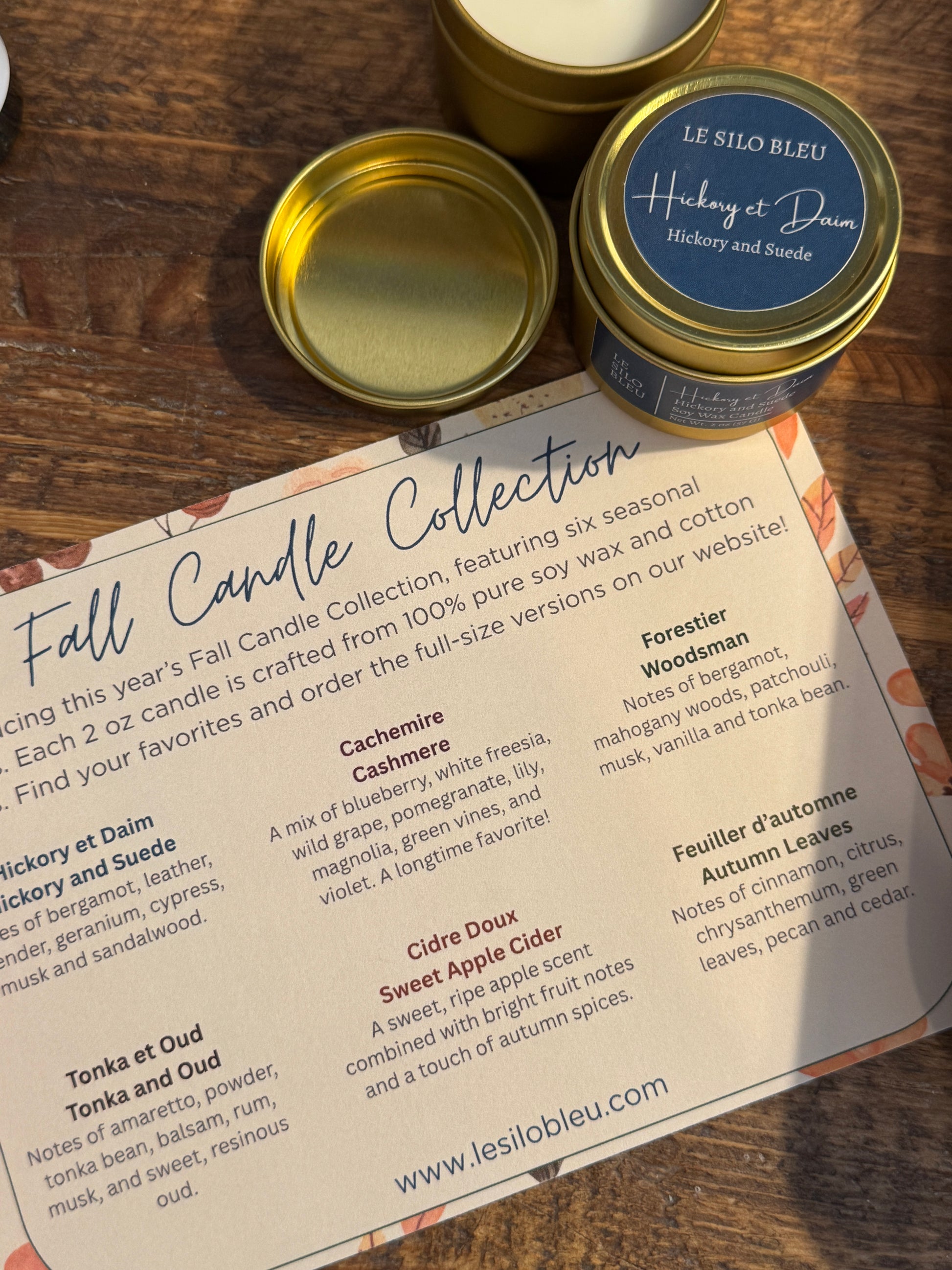 Three small candles with gold lids on a wooden surface next to a product description sheet.