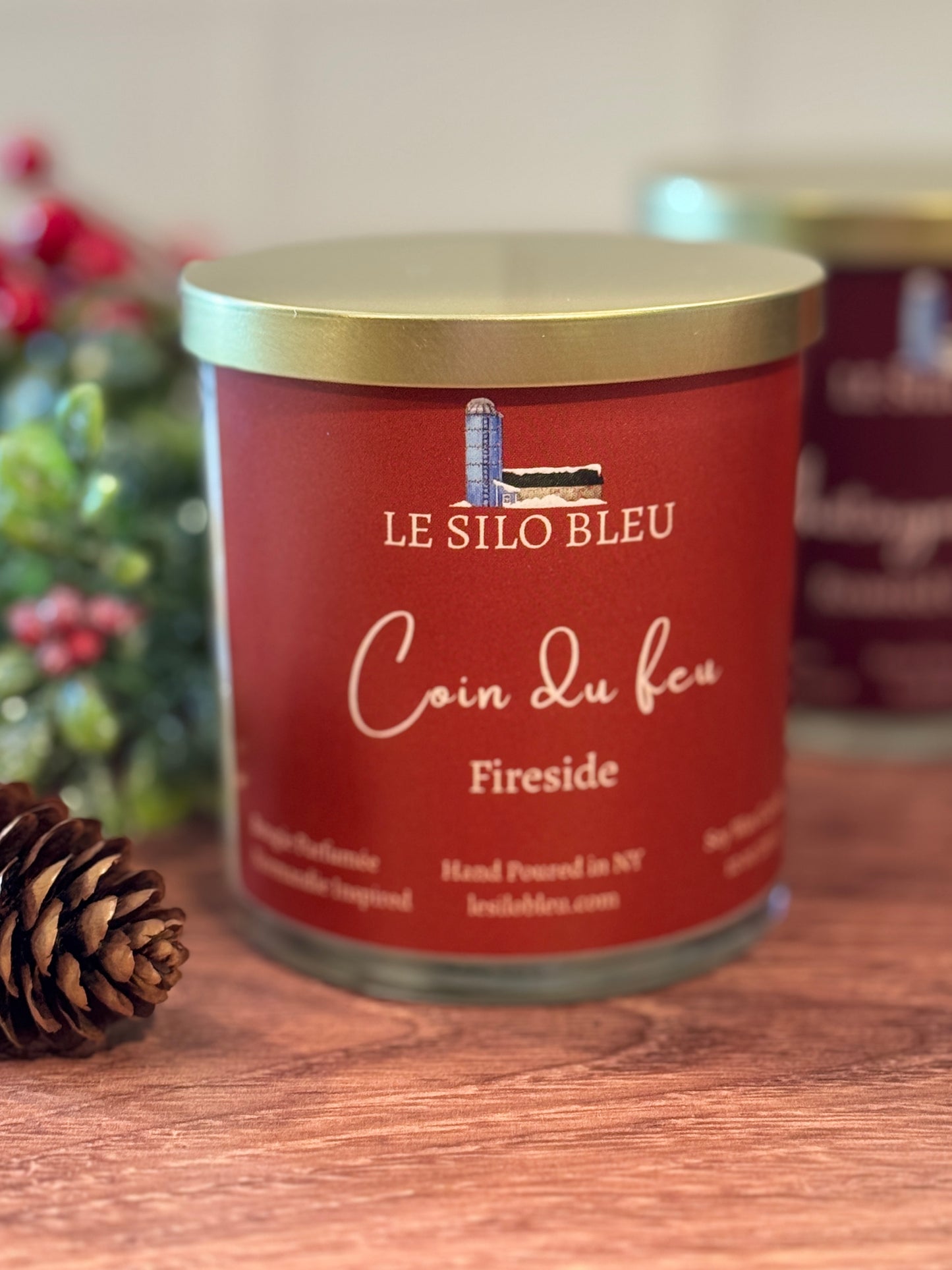 Candle jar labeled 'Le Silo Bleu Coin du feu Fireside' on a wooden surface with a pine cone.