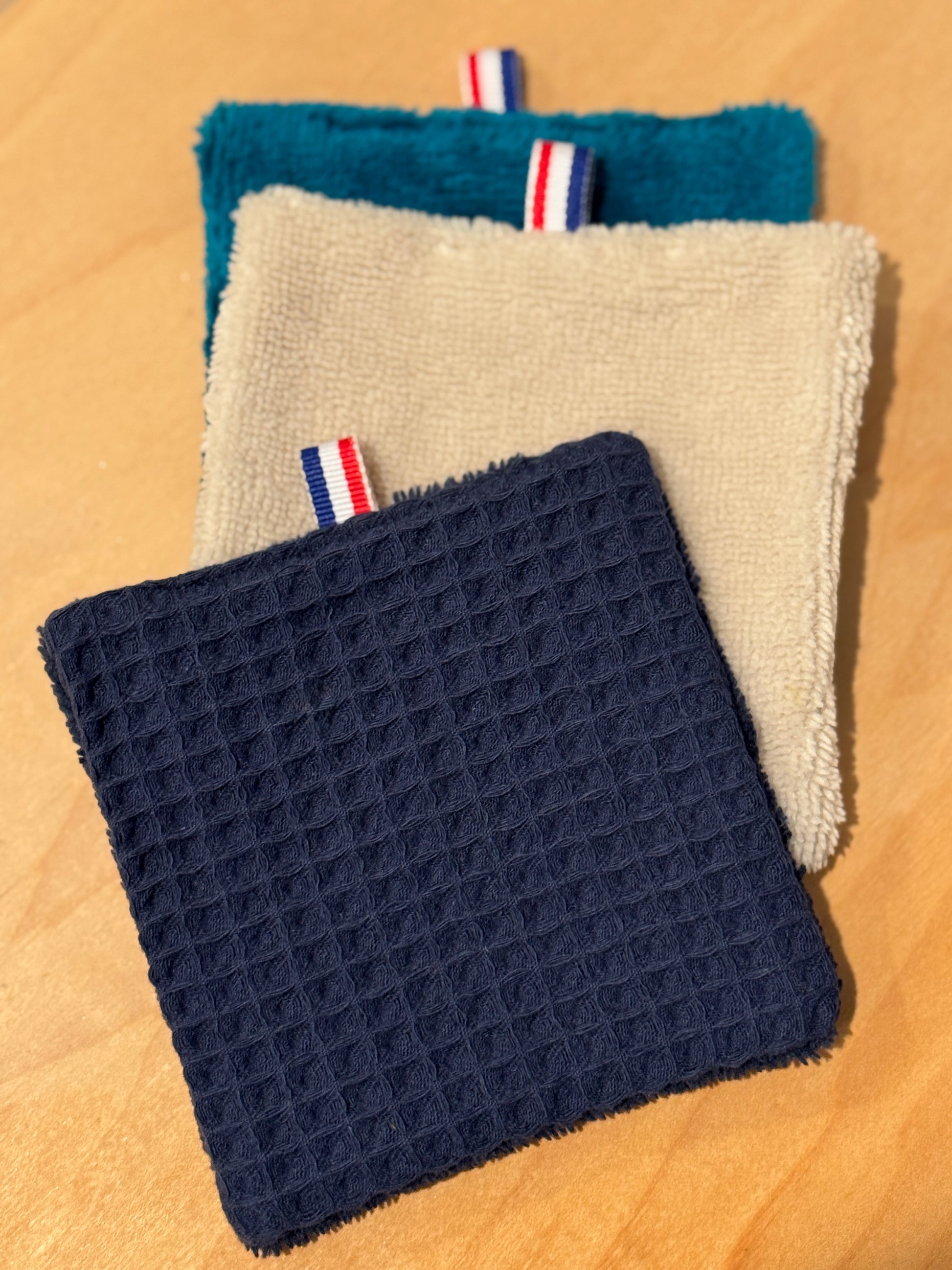 Three textured cotton cosmetic pads in blue, beige, and navy on a wooden surface. This shows both sides of the pads with terry on one side and cotton honeycomb patterned on the other.