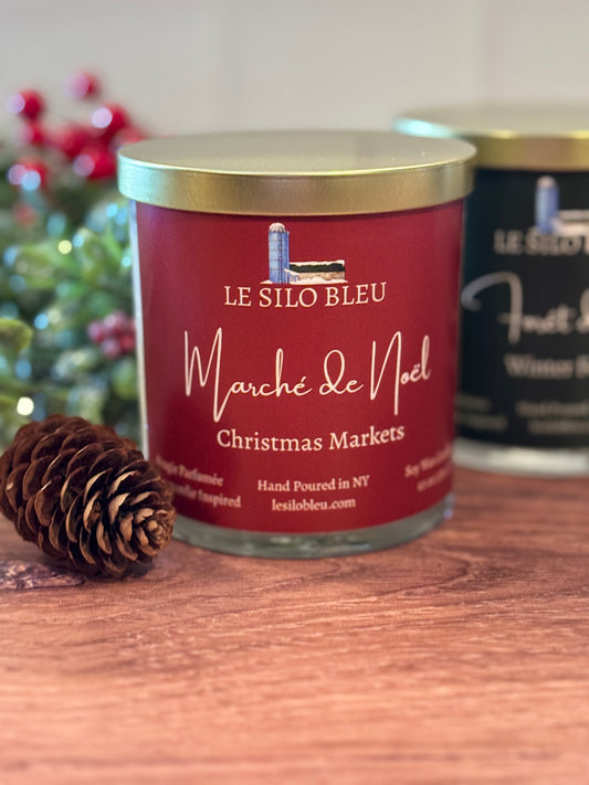 Red candle jar with gold lid labeled 'Le Silo Bleu Marché de Noel Christmas Markets' on a wooden surface.