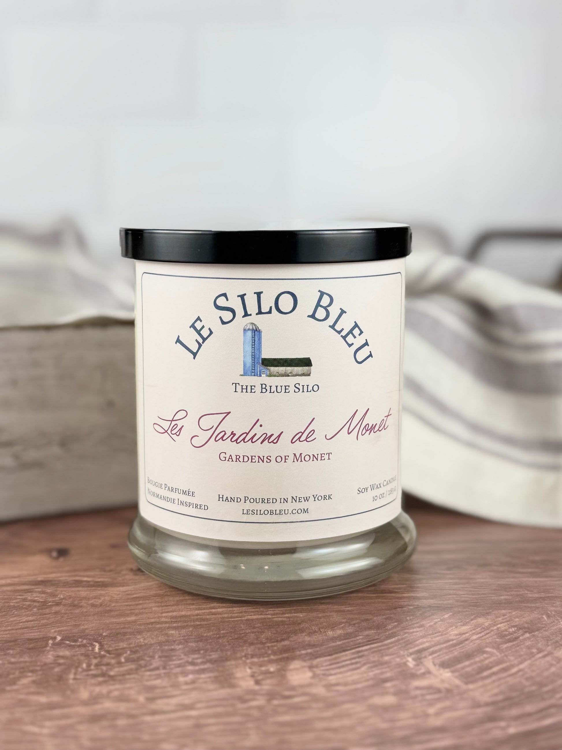 A large hand poured unlit soy wax candle in a glass jar labeled "Les Jardins de Monet - Gardens of Monet" sitting on a wooden tabletop with a black lid on the jar. In the background you can see a white linen towel with lavender stripes in an antique box.