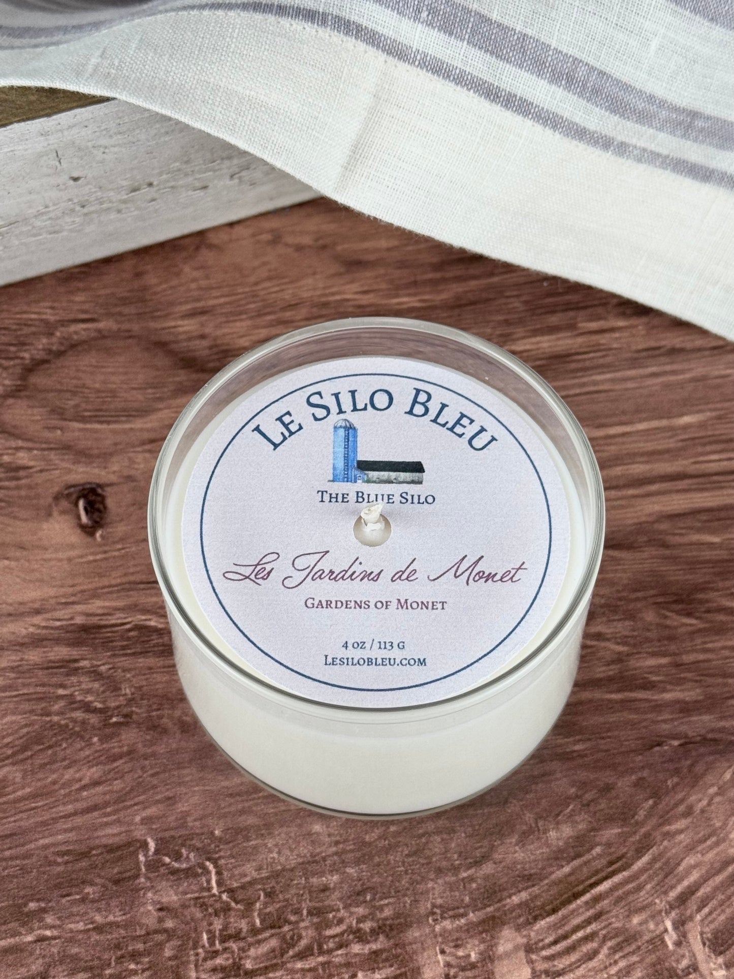 A small hand poured unlit soy wax candle in a glass jar labeled "Les Jardins de Monet - Gardens of Monet" sitting on a wooden tabletop. In the background there is a wooden antique box with a white and lavender striped linen towel. 