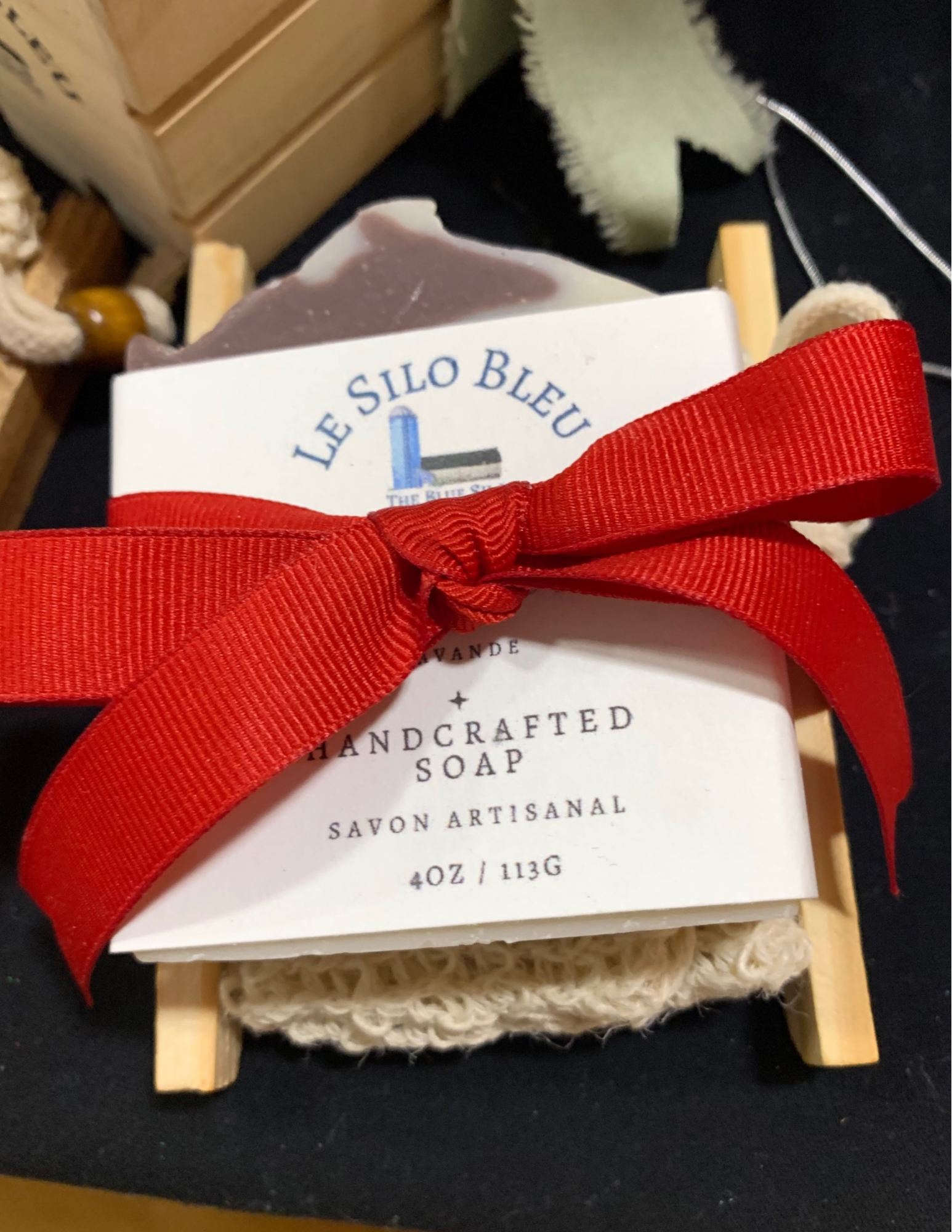 A bar of our handmade soap with a soap saver bag all tied together with a red ribbon on a wooden bamboo soap tray. This gift set is on top of a black tablecloth with a crate of soap and another gift bundle visible in the corners of this picture. 