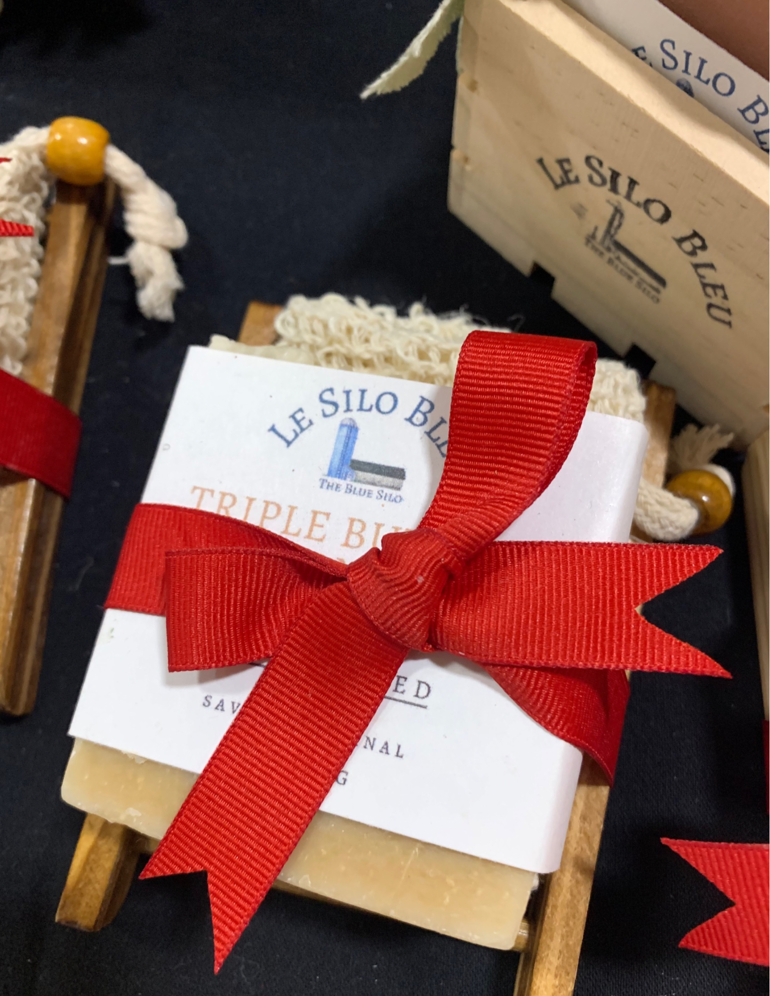 A bar of our handmade soap with a soap saver bag all tied together with a red ribbon  on a wooden bamboo soap tray. This gift set is on top of a black tablecloth with a crate of soap and another gift bundle visible in the corners of this picture. 