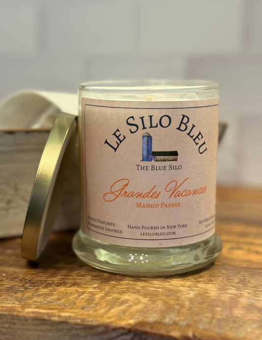 Grandes Vacanes, a mango papaya  scented candle with gold lid to the side. Candle is sitting on top of a wood table with an antique box and linen cloth in the background.