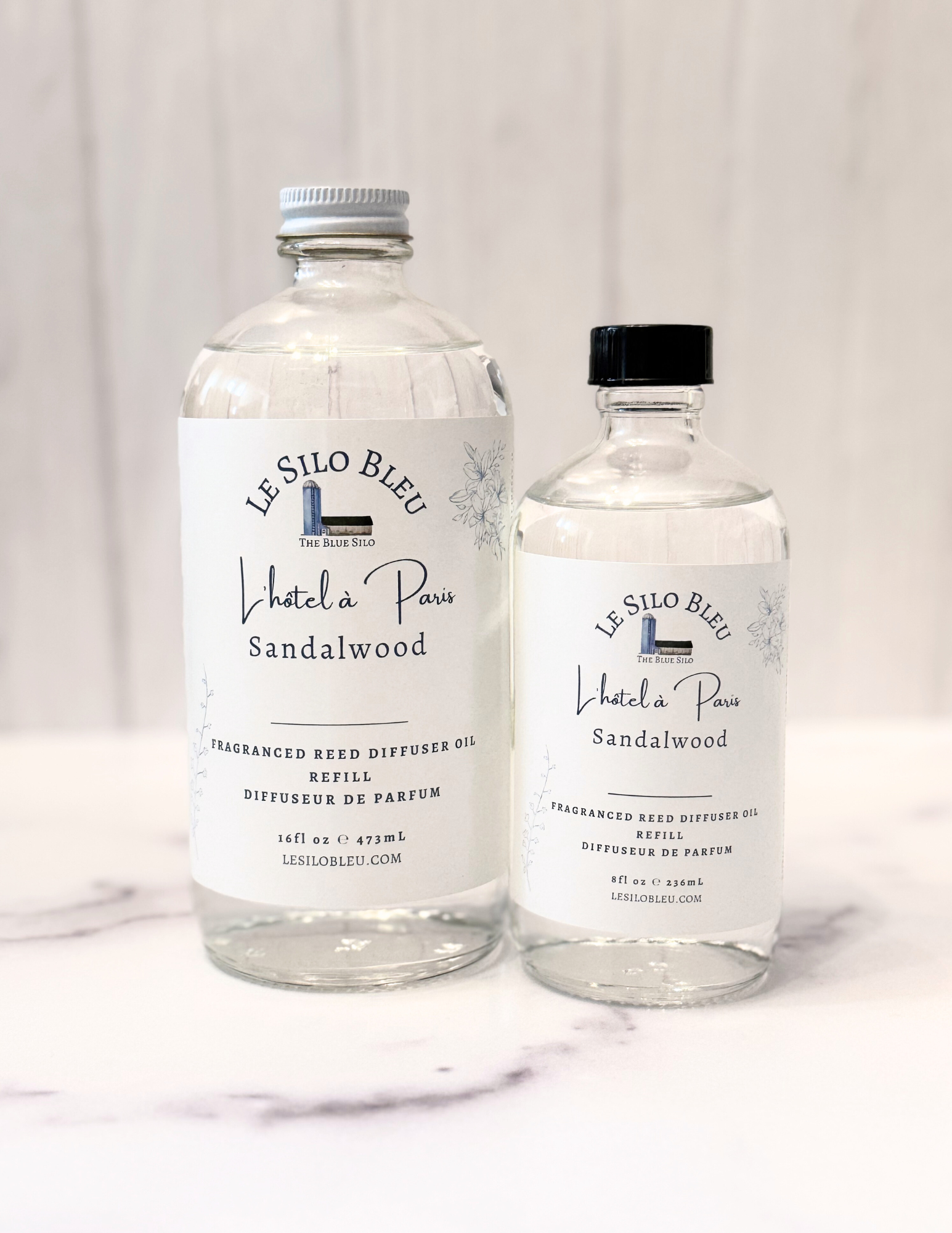 Hotel in Paris Reed Diffuser Refill bottles in 8oz and 16oz glass bottles are shown sitting on a white marble table with a white wood background. The cap for the large bottle is white and the cap for the small bottle is black. 
