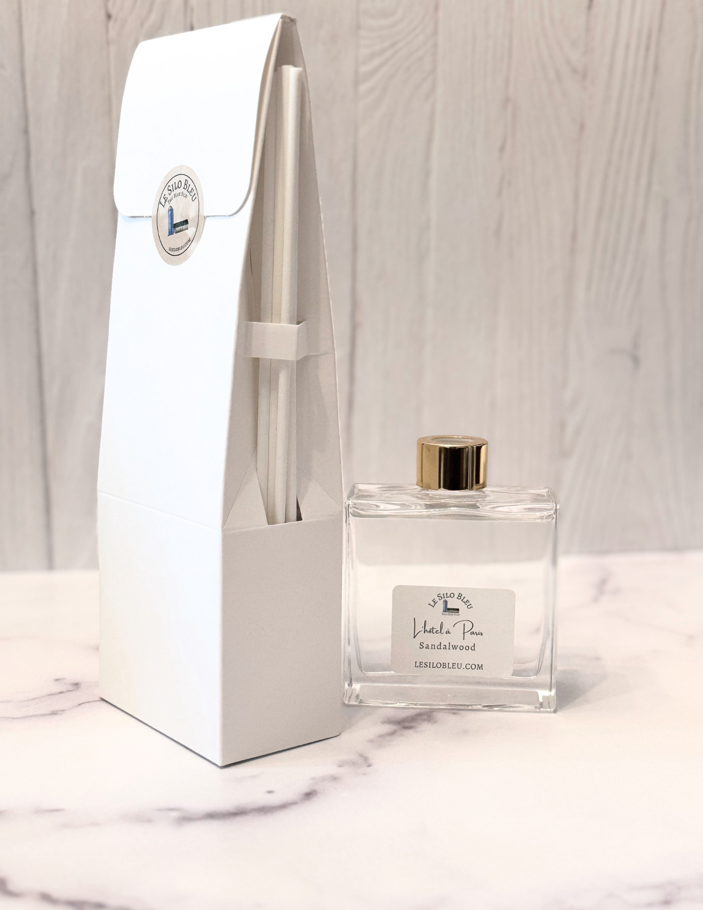 Reed diffuser bottle with gold cap and white label next to its packaging on a marble surface.