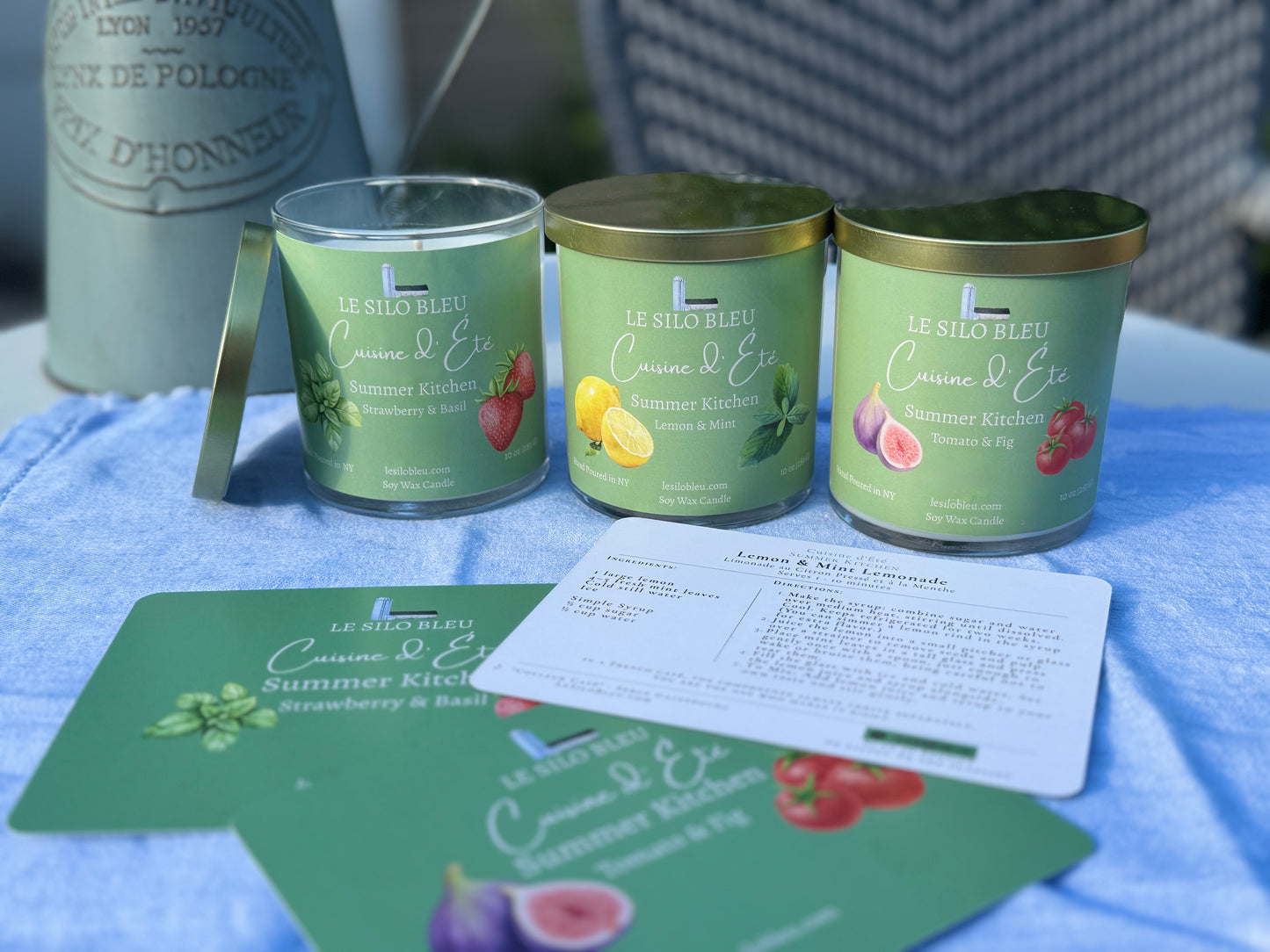 Three Kitchen Garden candles with gold lids on a blue linen surface, accompanied by matching recipe  cards.