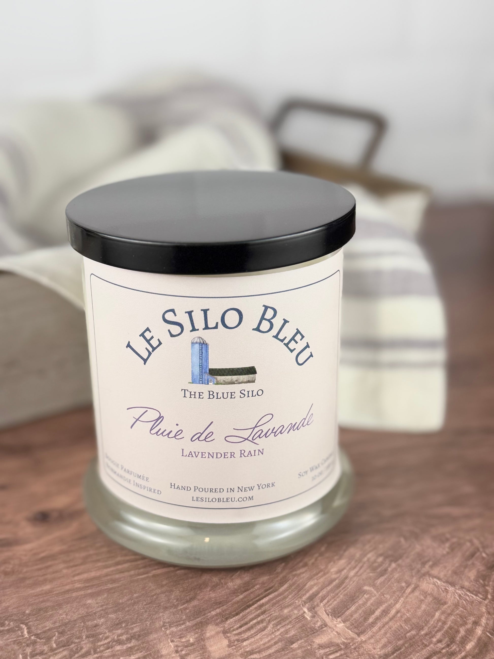 A handpoured unlit soy wax candle in a glass jar labeled "Pluie de Lavender - Lavender Rain" sitting on a wooden table top with a black lid on  the jar. In the background  there is a white linen towel with lavender stripes in an antique box with a metal handle. 