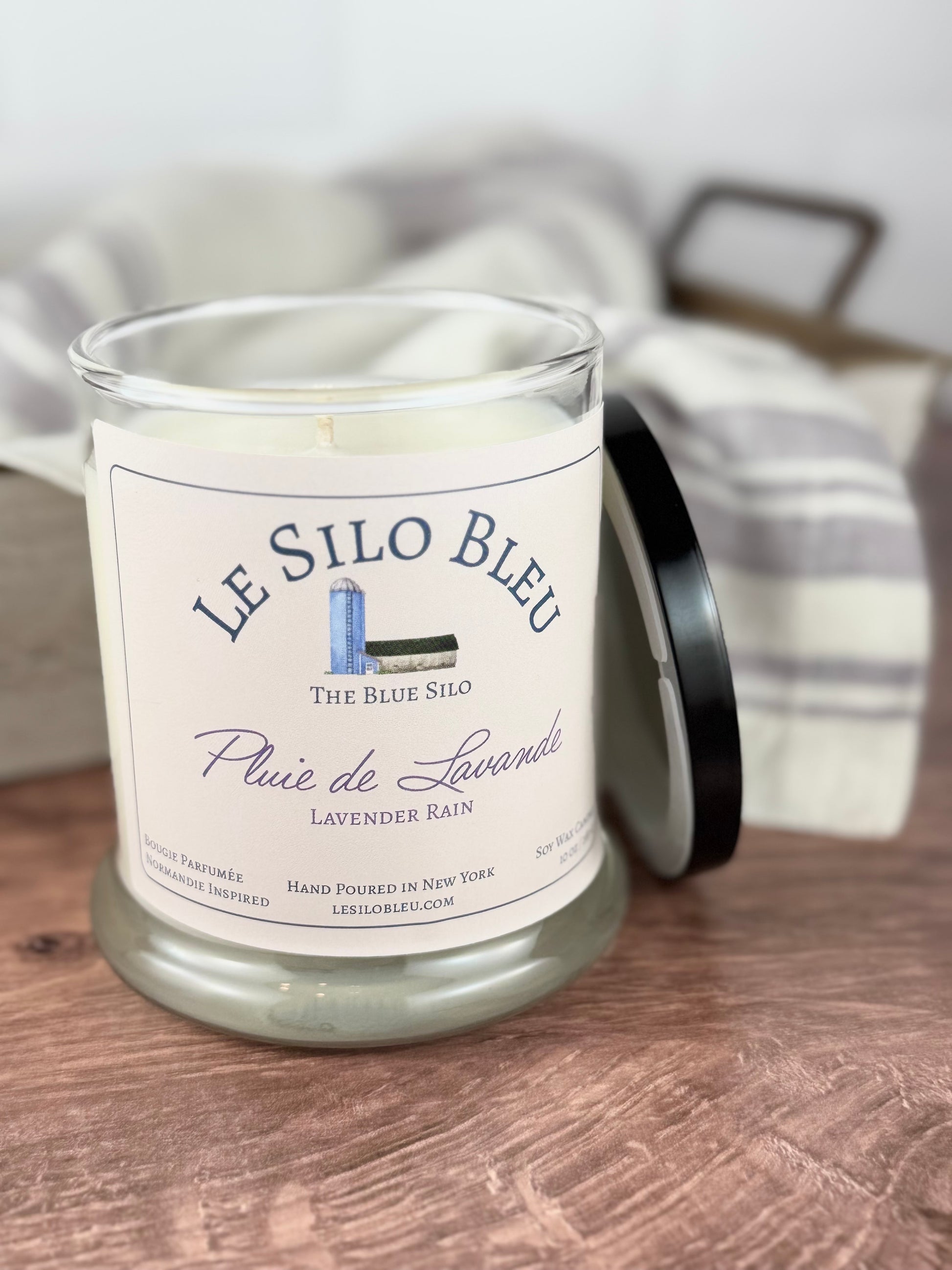 A handpoured unlit soy wax candle in a glass jar labeled "Pluie de Lavande- Lavender Rain" sitting on a wooden table top with a black lid next to the jar. In the background there is a white linen towel with lavender stripes in an antique box with a metal handle.