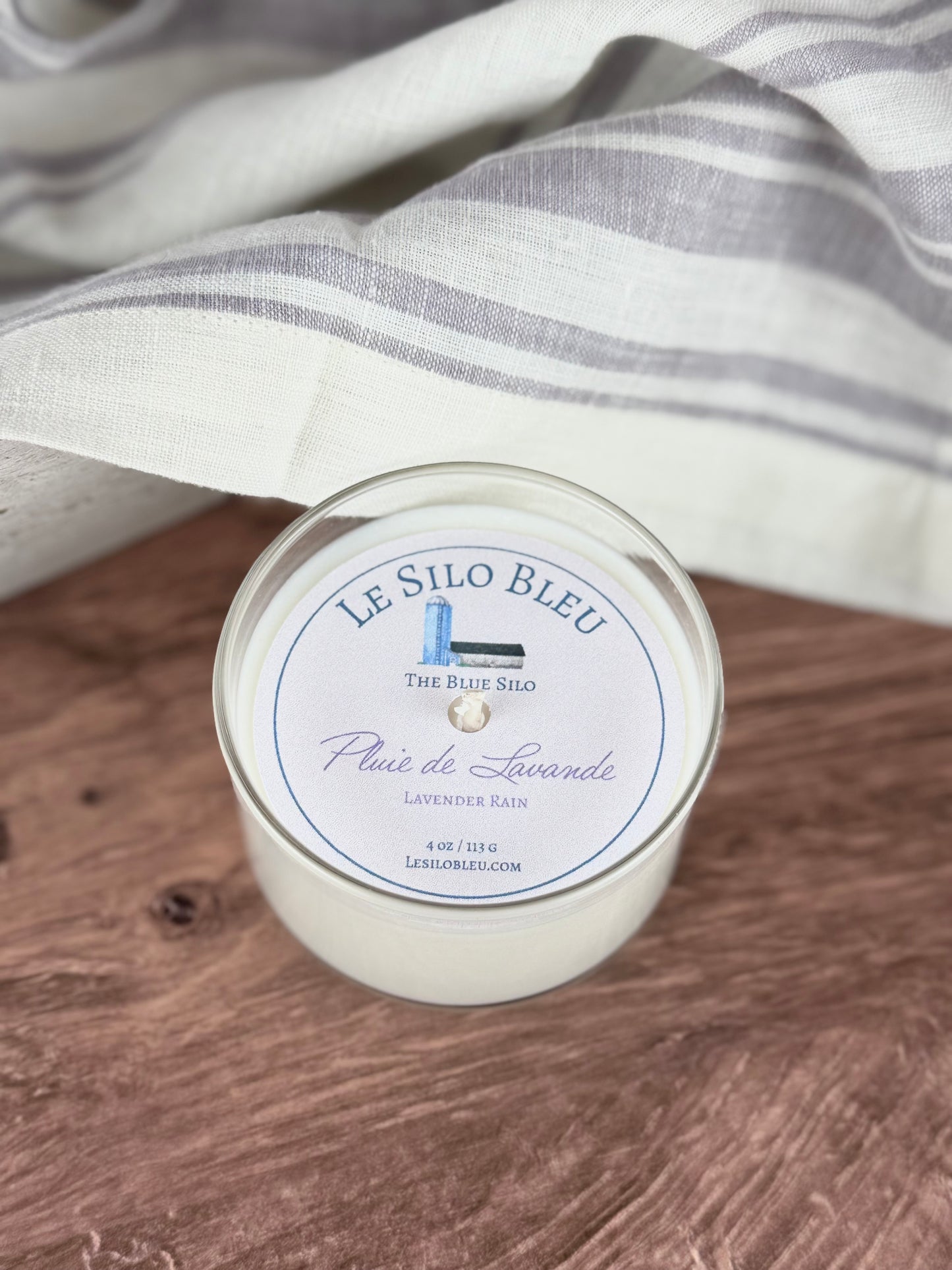 A small handpoured unlit soy wax candle in a glass jar labeled "Pluie de Lavande- Lavender Rain" sitting on a wooden table top. In the background there is a white linen towel with lavender stripes in an antique box.