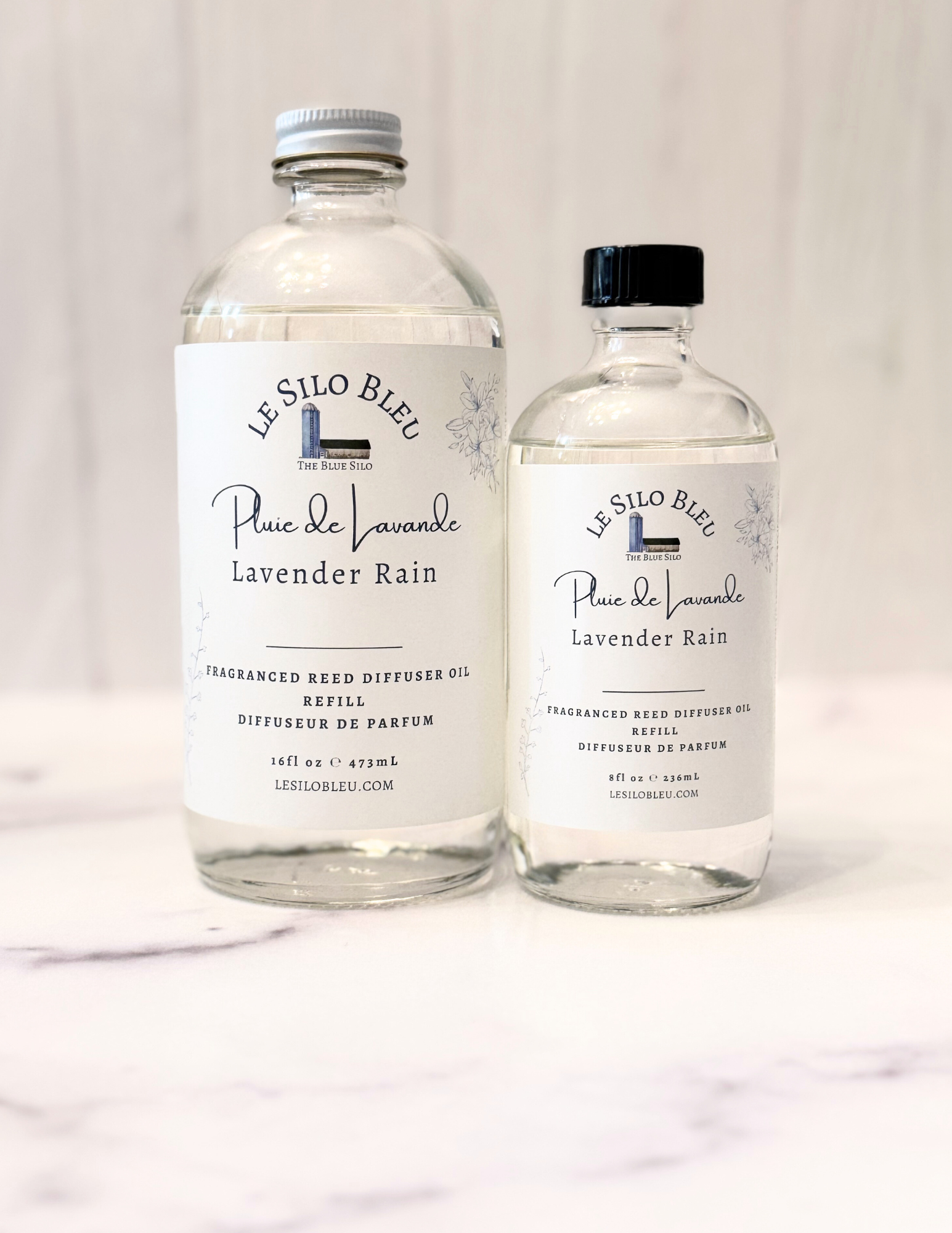 Lavender Rain Reed Diffuser Refill bottles in 8oz and 16oz glass bottles are shown sitting on a white marble table with a white wood background. The cap for the large bottle is white and the cap for the small bottle is black. 