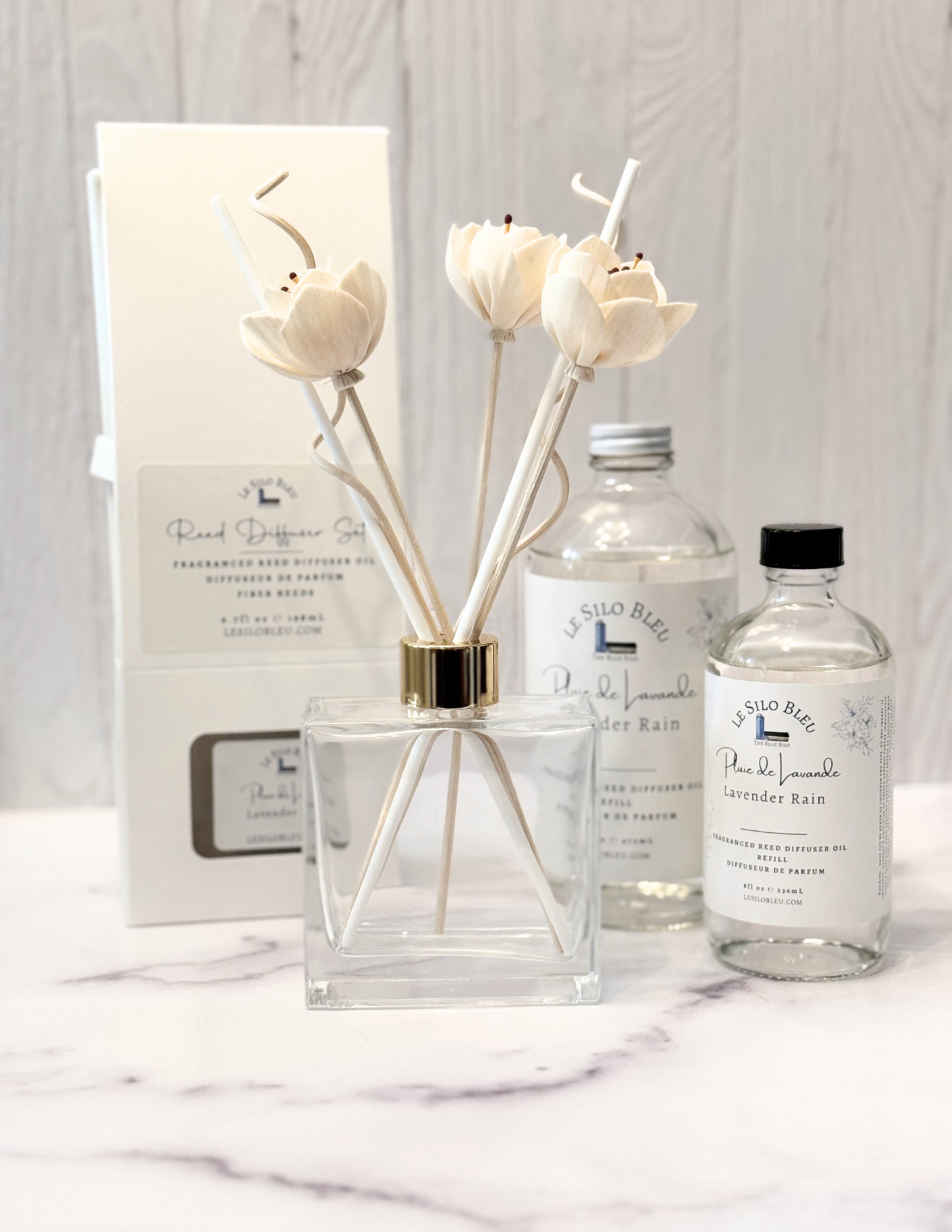 Lavender Rain Scented Reed Diffuser set and collection showcasing the optional floral reeds along with the included white reeds and additional for purchase bottles of 8oz or 16oz fragrance refills.  The whole scent collection is sitting on a white marble table with a white wood background. 