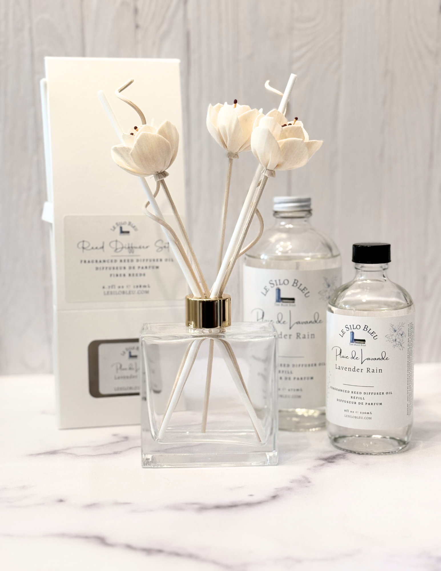 Lavender Rain Scented Reed Diffuser set and collection showcasing the optional floral reeds along with the included white reeds and additional for purchase bottles of 8oz or 16oz fragrance refills.  The whole scent collection is sitting on a white marble table with a white wood background. 