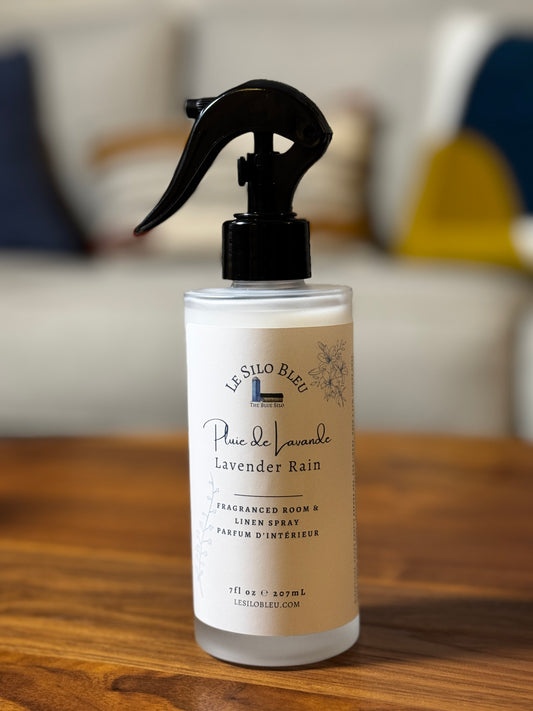 A frosted glass room spray bottle with a black trigger sprayer, labeled 'Le Silo Bleu' and 'Lavender Rain', sitting on a wooden table with a blurred living room background.