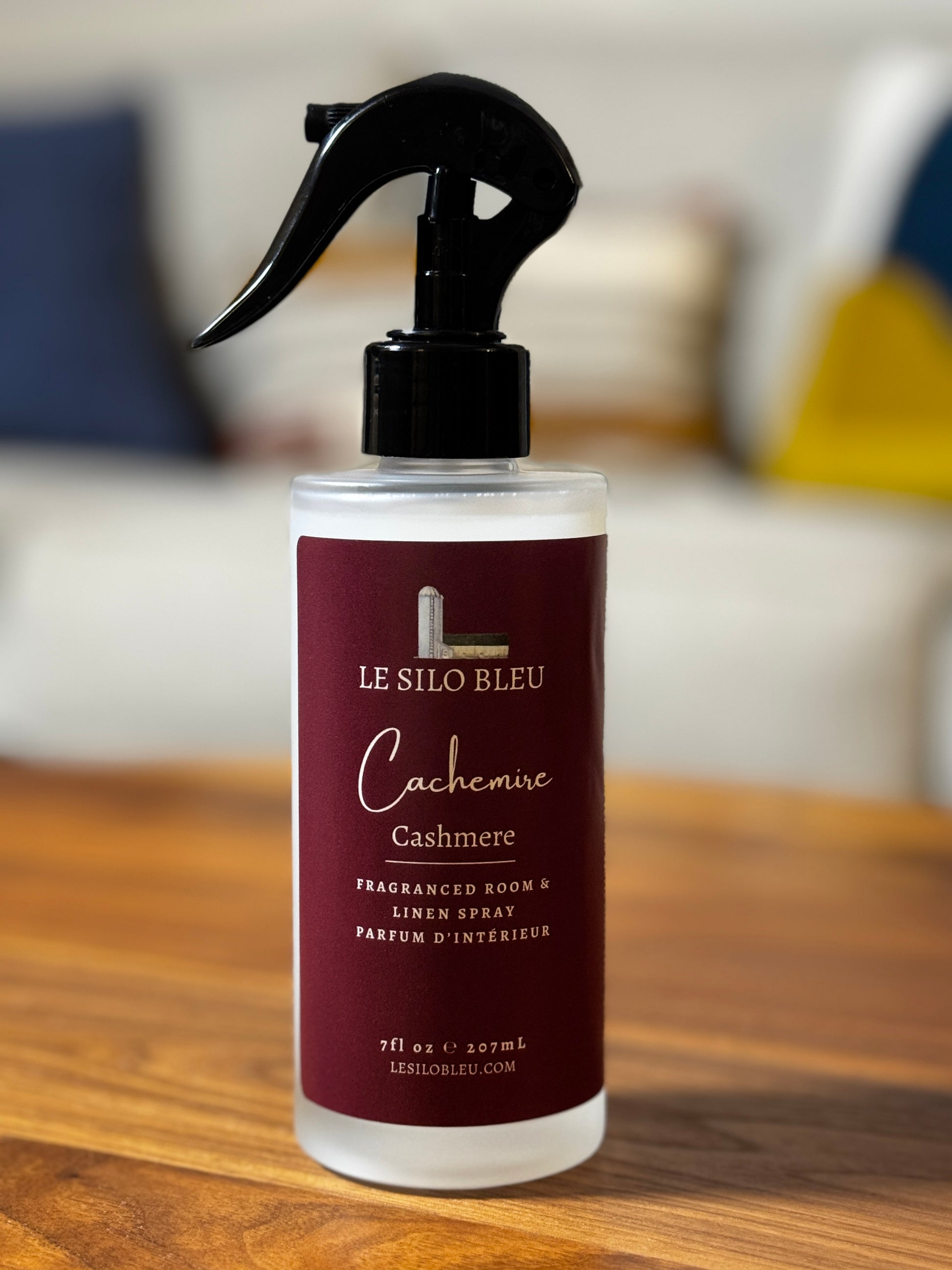 A bottle of Le Silo Bleu Cashmere fragranced room and linen spray, featuring a deep burgundy label and a black trigger spray top, resting on a wooden table with a blurred living room background.