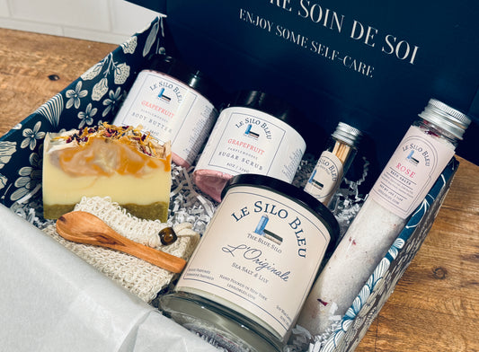 Le Silo Bleu self-care products with candle, bathsalts tube, handcrafted soap, and body butter sugar scrub set in a branded box sitting on a wooden surface.