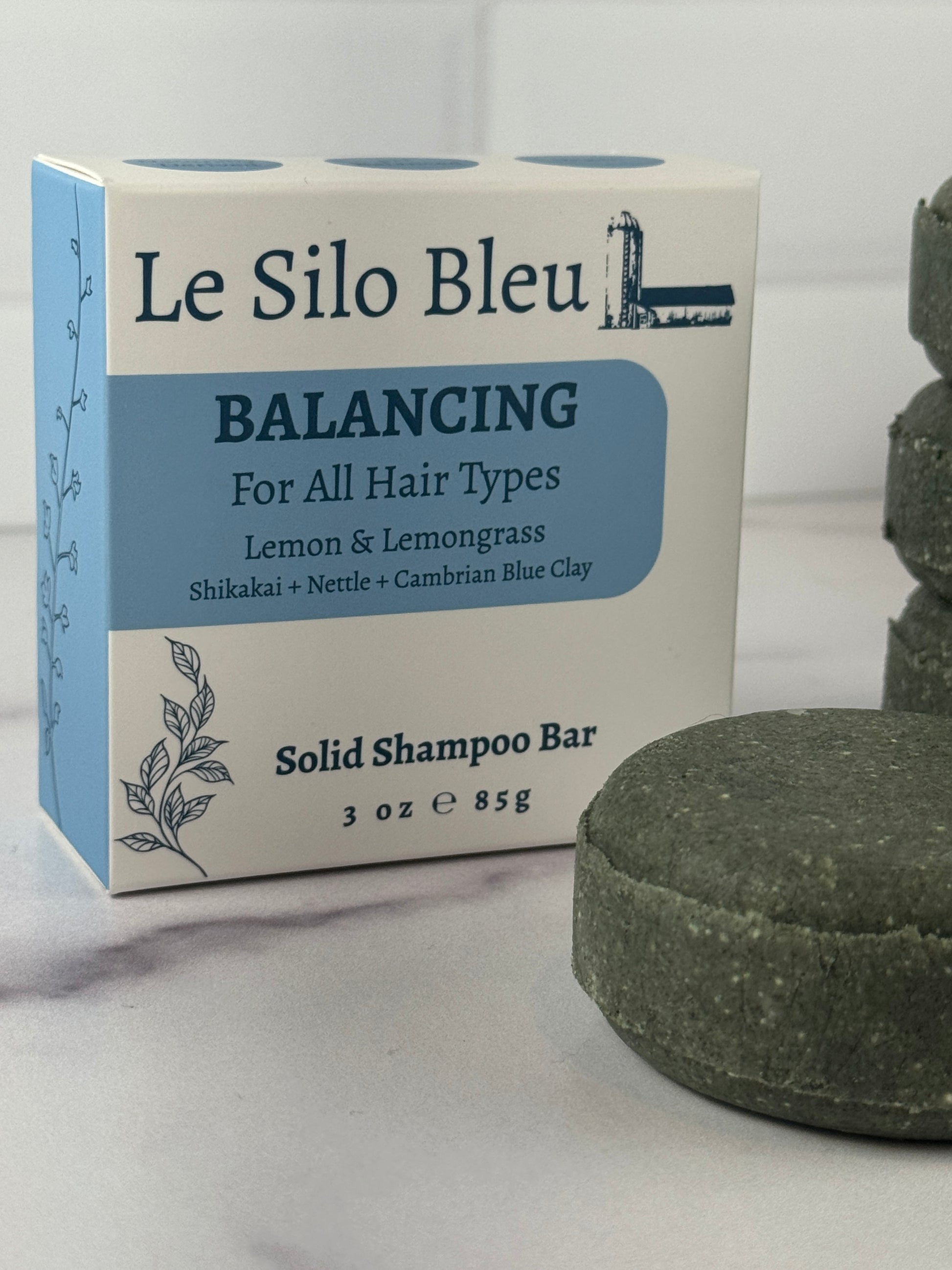 A dark green Le Silo Bleu Balancing solid shampoo bar with packaging sitting on a white marble surface with a white tiled background. 