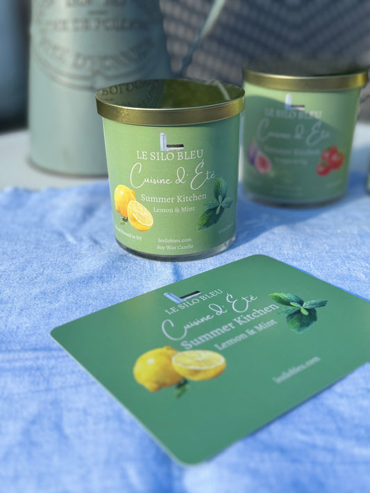 Summer Kitchen Lemon Mint Candle with a green label on a blue surface, surrounded by similar candles and a pitcher of flowers in the background. 
