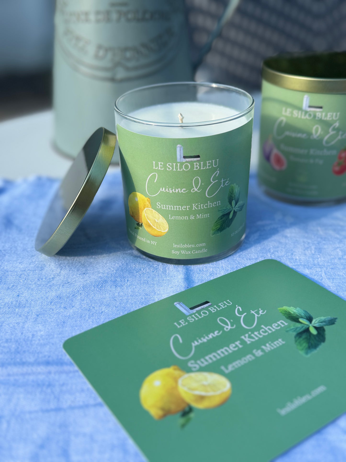 Summer Kitchen Lemon Mint Candle with a green label on a blue surface, surrounded by similar candles and a pitcher of flowers in the background. 