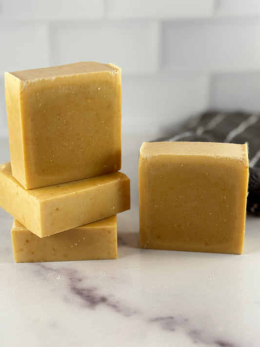 Four  bars of handmade turmeric soap in a warm golden-yellow color, with three stacked on the left and one standing on the right, set on a white marble surface with a blurred white subway tile background.