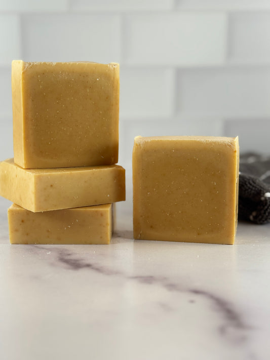 Three bars of light turmeric handmade soap in a warm golden-yellow color, with two stacked on the left and one standing on the right, set on a white marble surface with a white subway tile background.