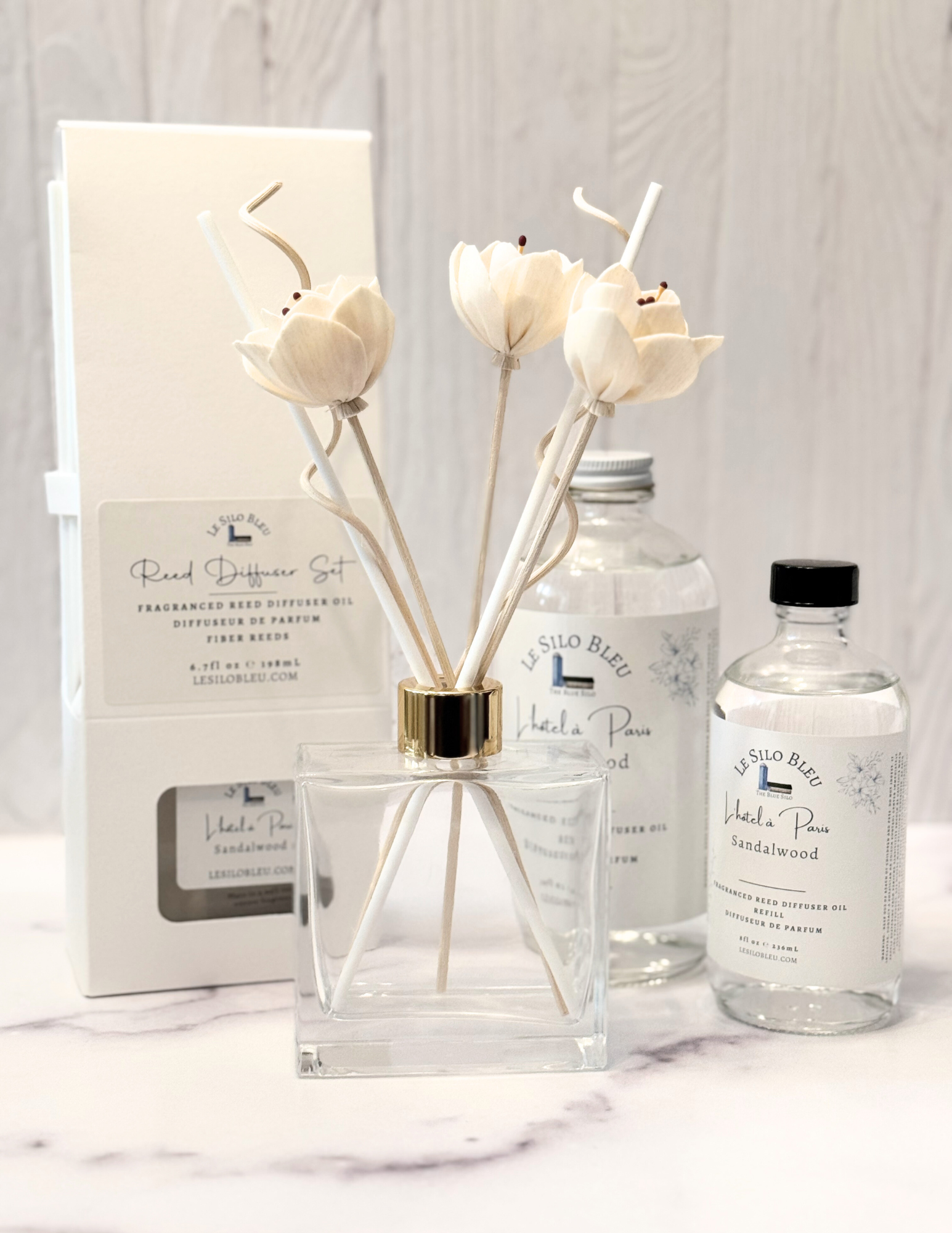 Reed Diffuser bottle showing the limited edition floral reed bundle surrounded by a collection of reed diffuser products sitting on a white marble table and a white wood background. 
