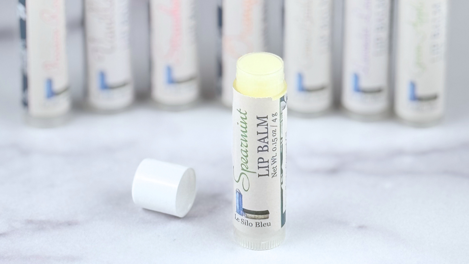 Tube of spearmint lip balm open in front of all the tubes on a white marble tabletop. 