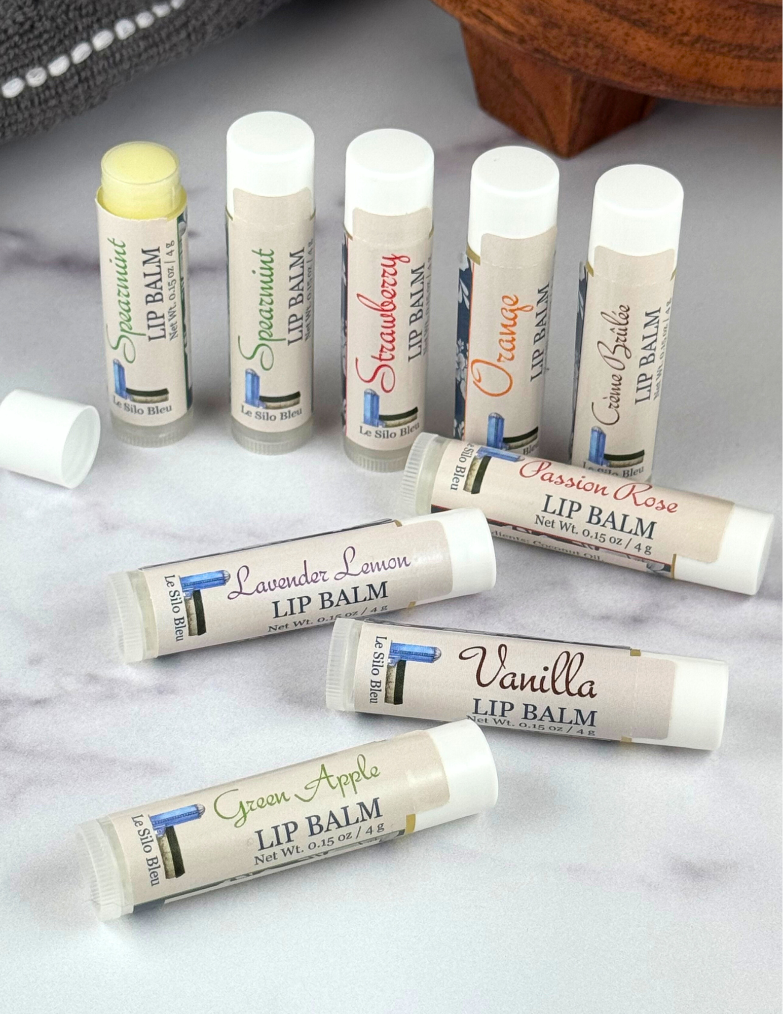 Lip Balm collection line up with one in back open all on a white marble table with a wood stand and grey towel in the background.