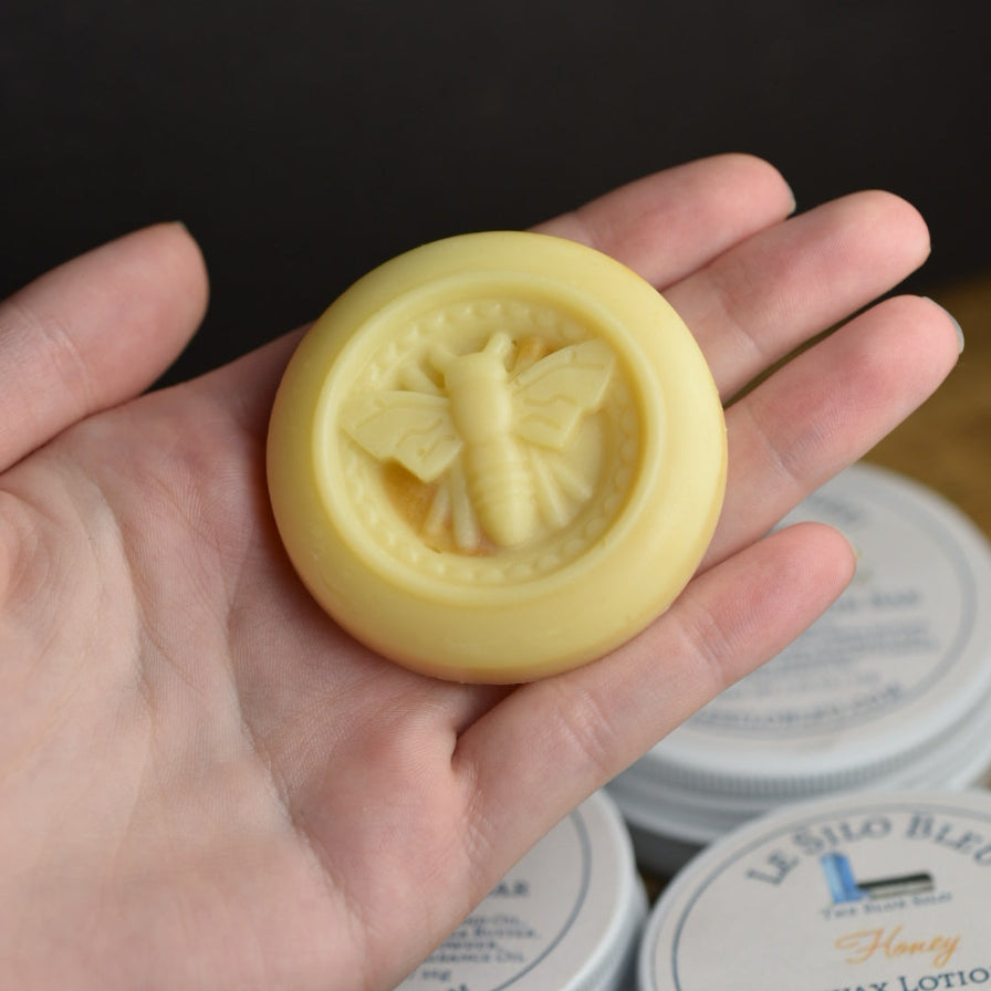 A hand holding a bee embossed lotion bar above a wooden table with three of the product tins visible. the backrgound is black. 